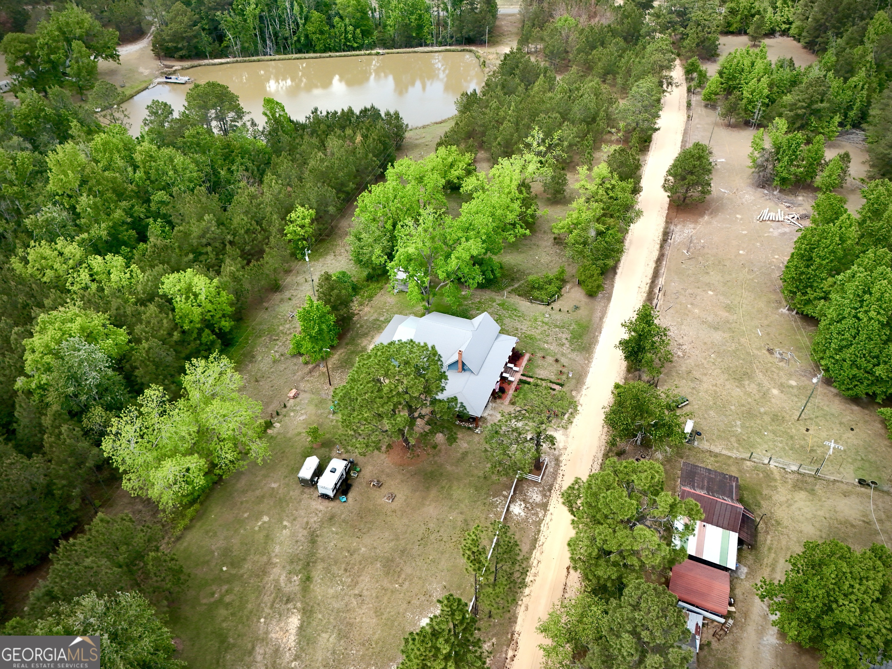 Aerial view of a rural home among trees, with a pond, outbuildings, and a dirt road.