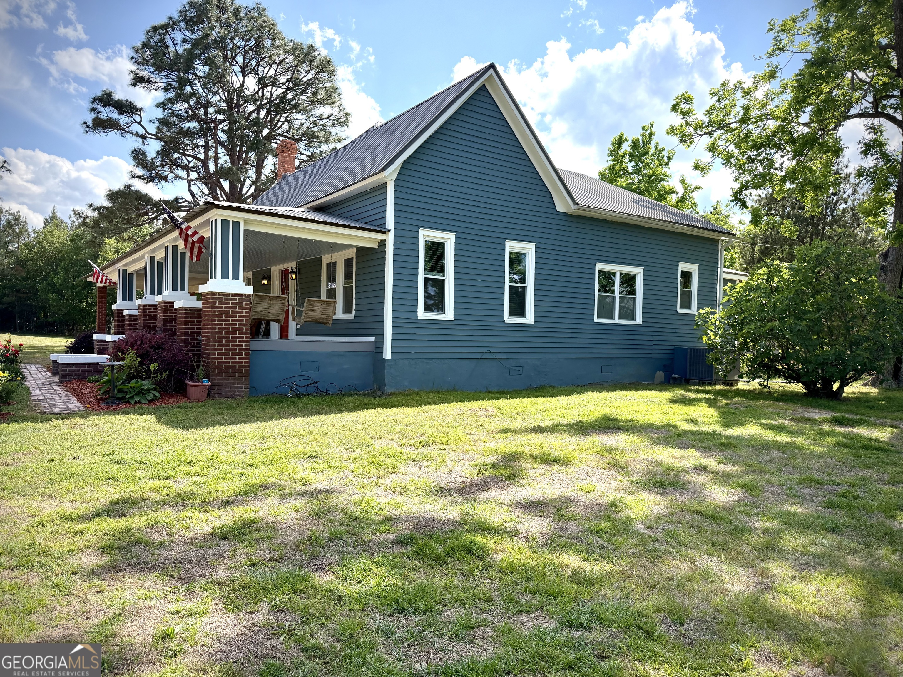 Blue-sided house with a brick-columned porch, metal roof, and broad grassy yard.