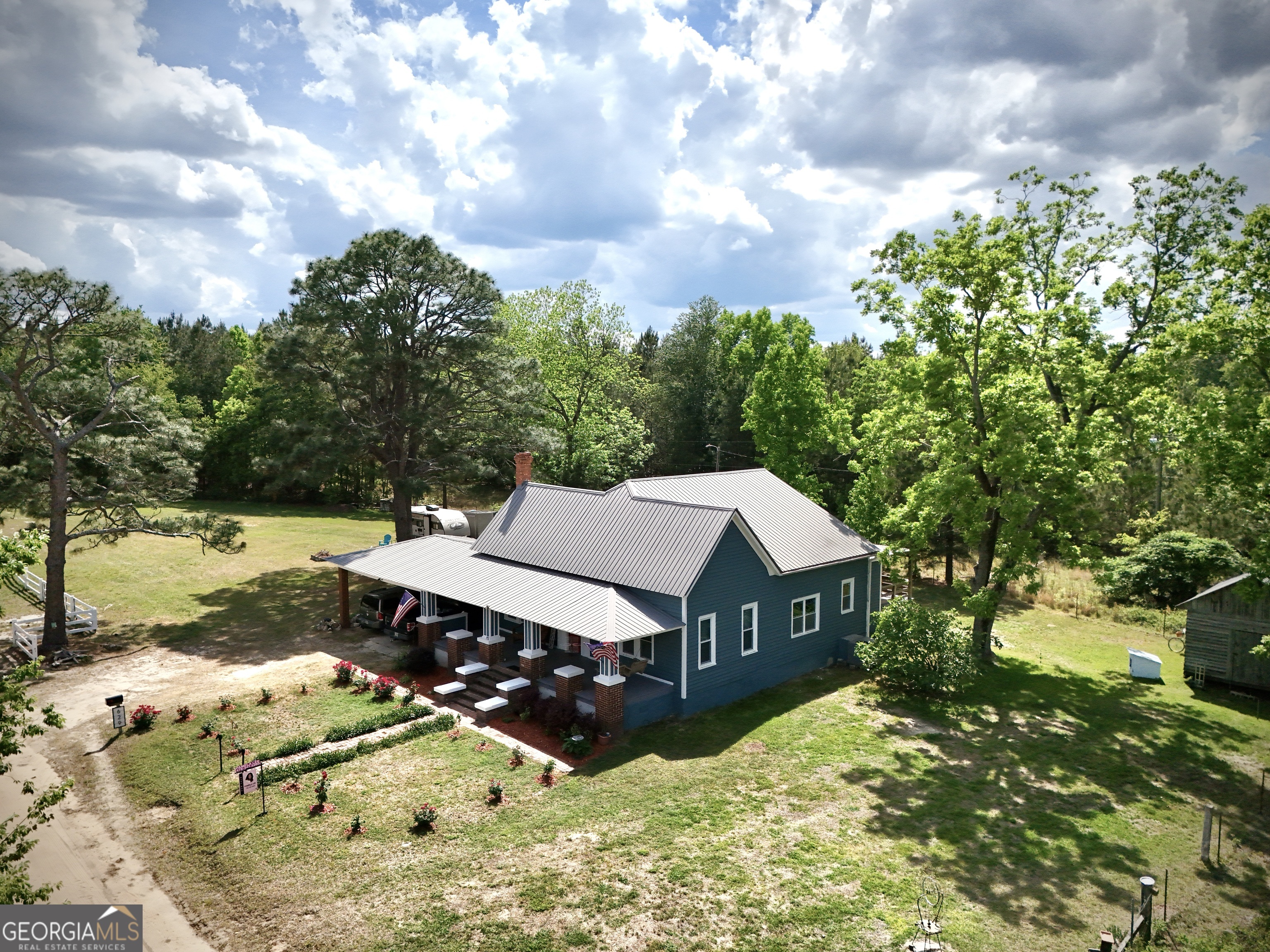 Aerial view of a blue house with metal roof, covered porch, and wooded lawn.