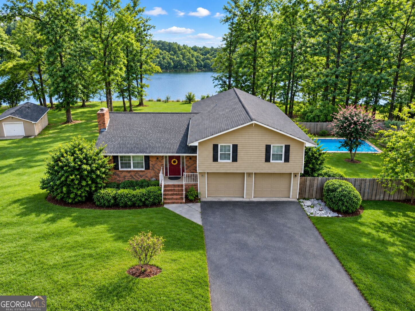 Aerial view of a brick-and-siding home with a pool beside a wooded lake.