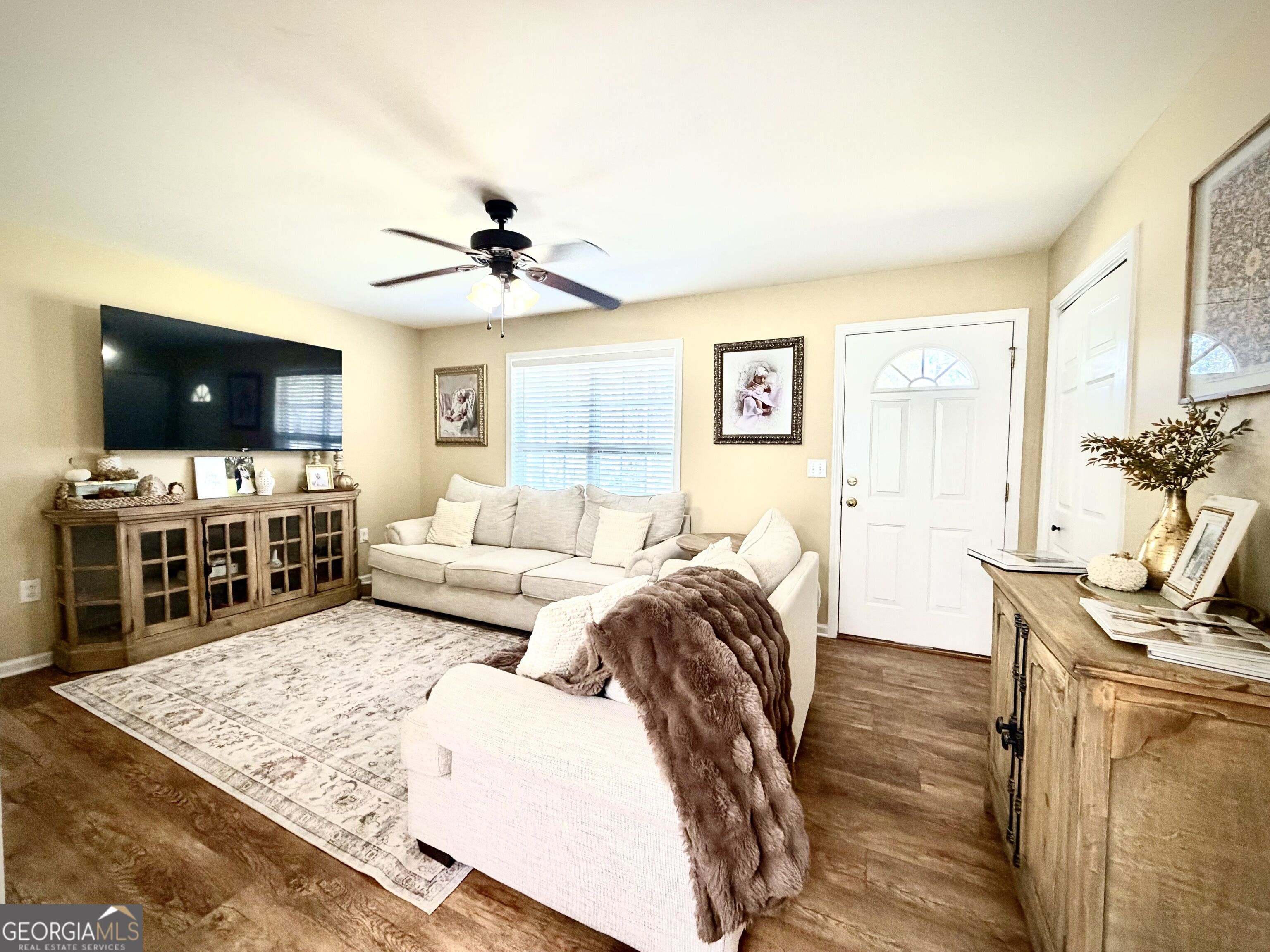 Living room with a light sectional, wall-mounted television, ceiling fan, and warm wood flooring.