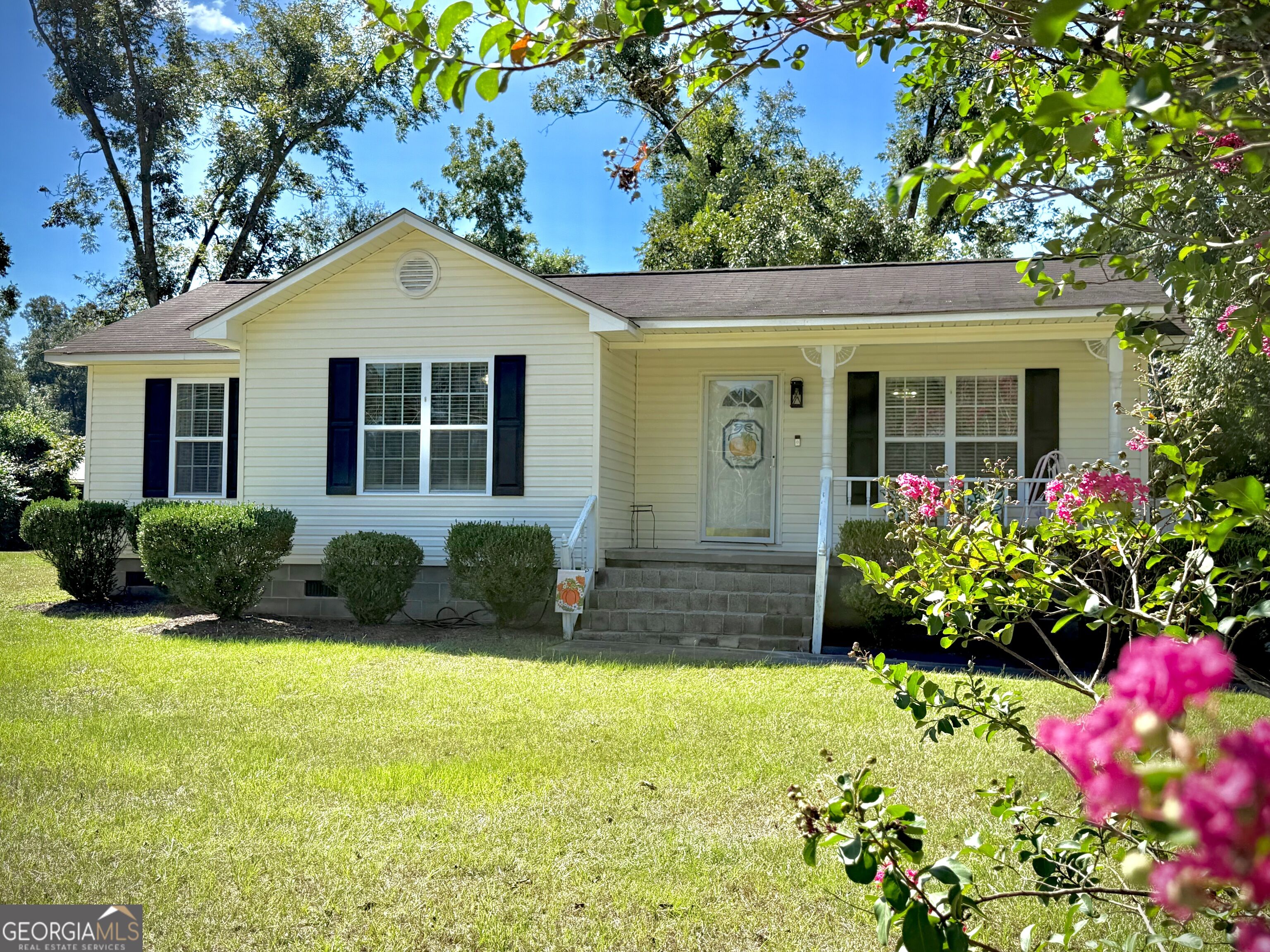 Cream-sided single-story house with dark shutters, a covered porch, and pink flowers beside the lawn.