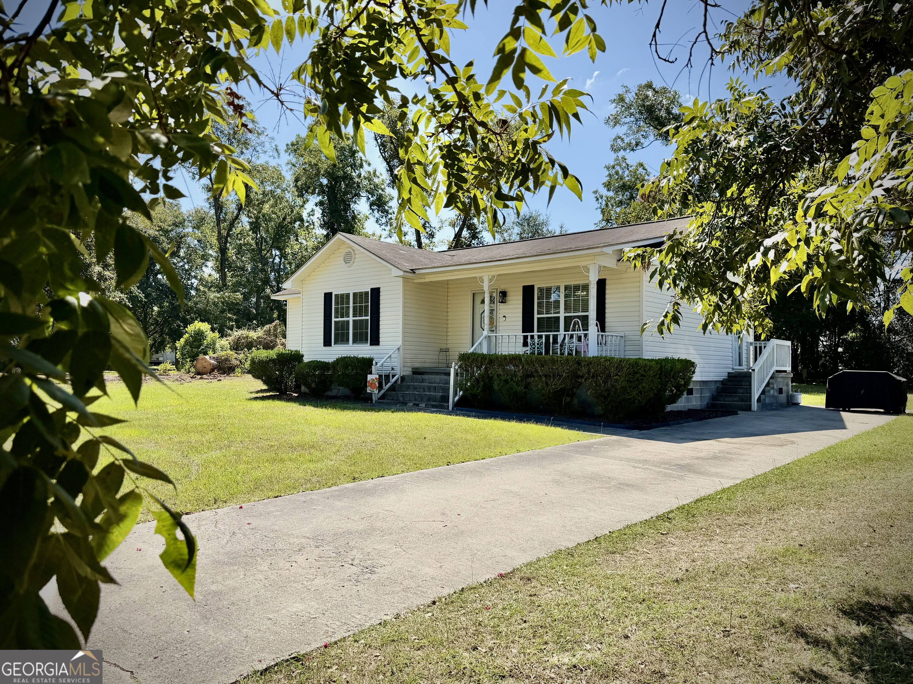 White single-story house with a covered porch and wide driveway, framed by leafy trees.