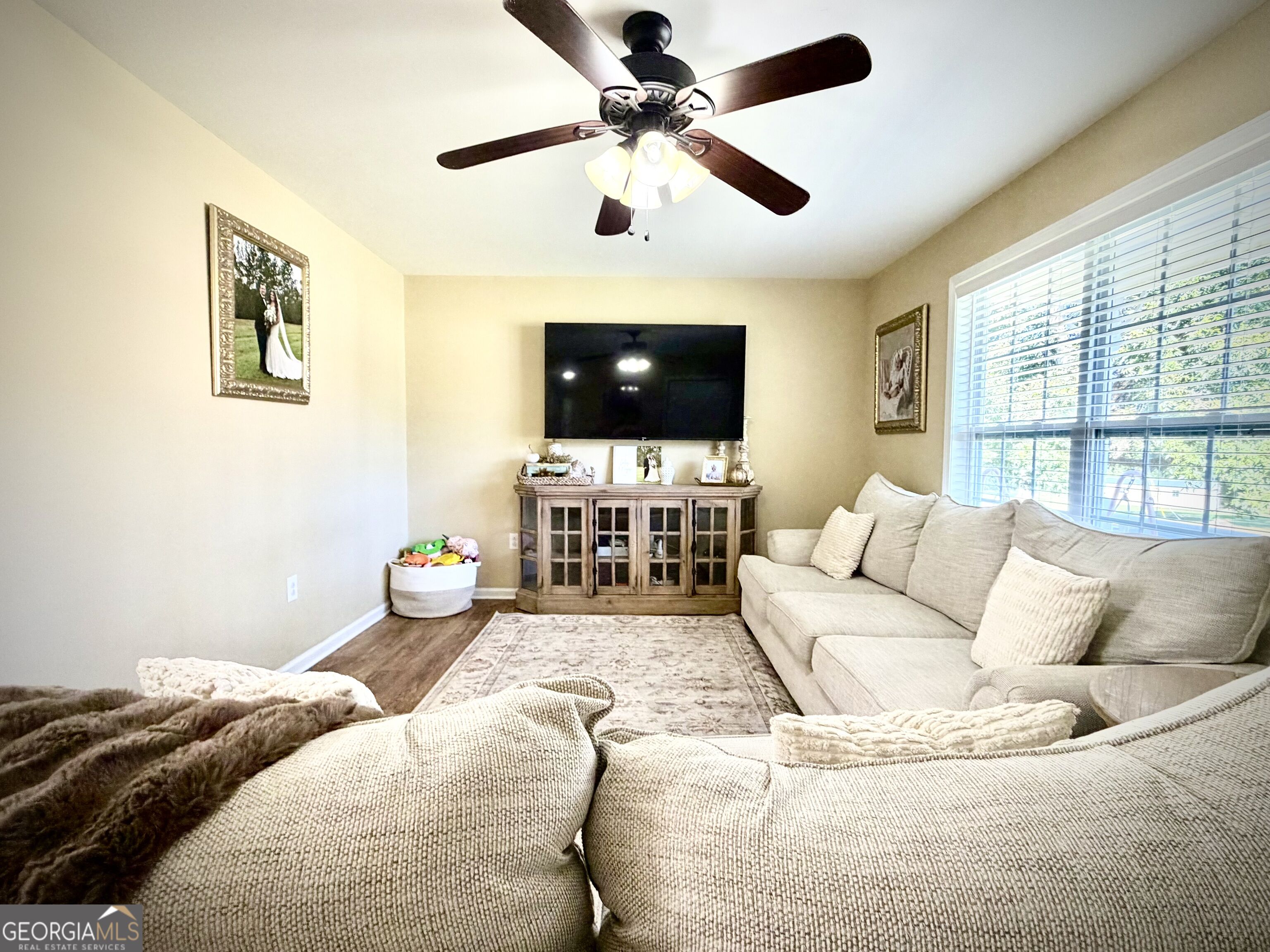 Furnished living room with a beige sectional, ceiling fan, TV console, and large window with blinds.