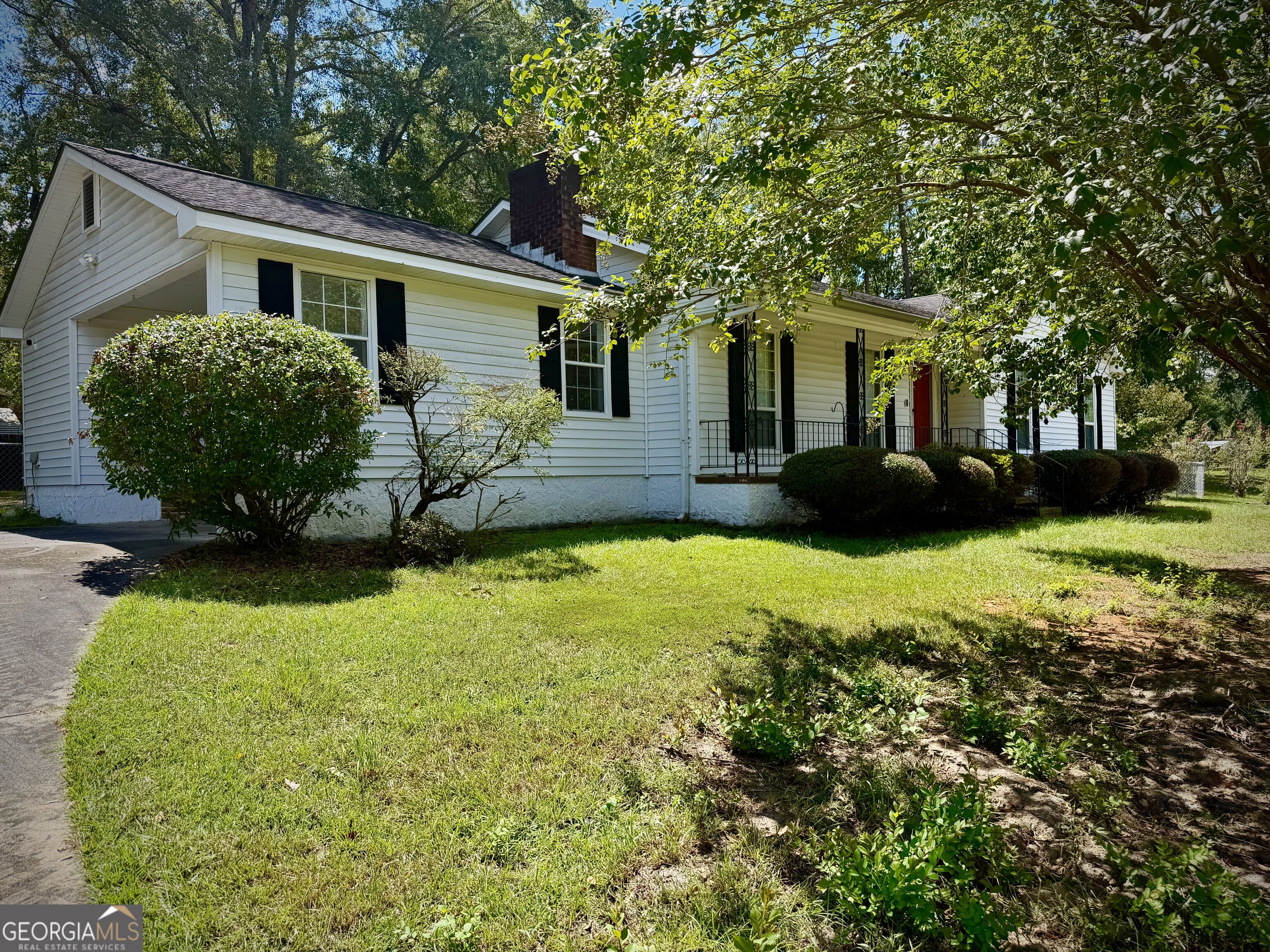 White single-story house with black shutters, red front door, covered porch, and leafy trees framing the lawn.