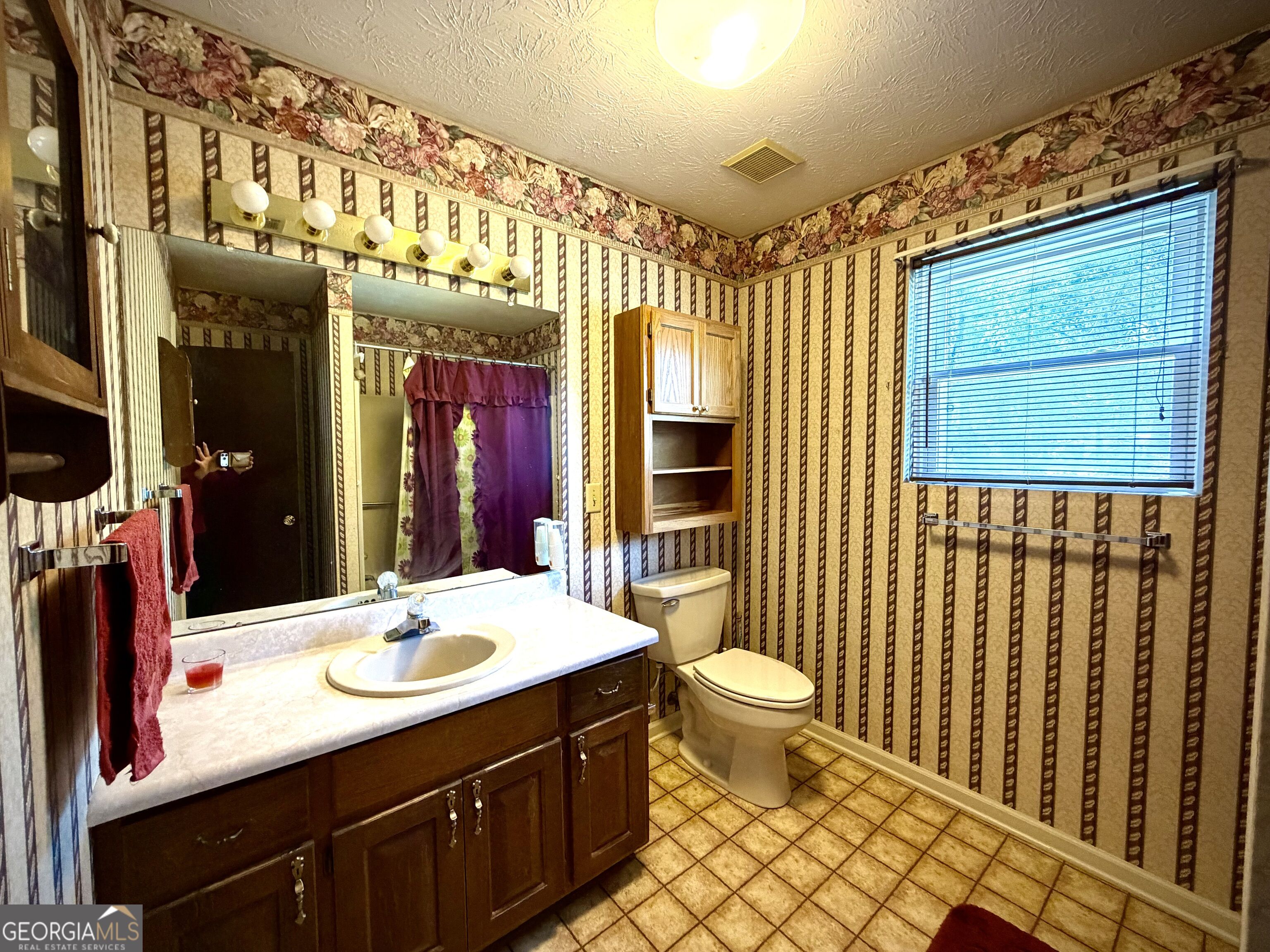 Bathroom with a dark wood vanity, patterned wallpaper, and a window with blinds.