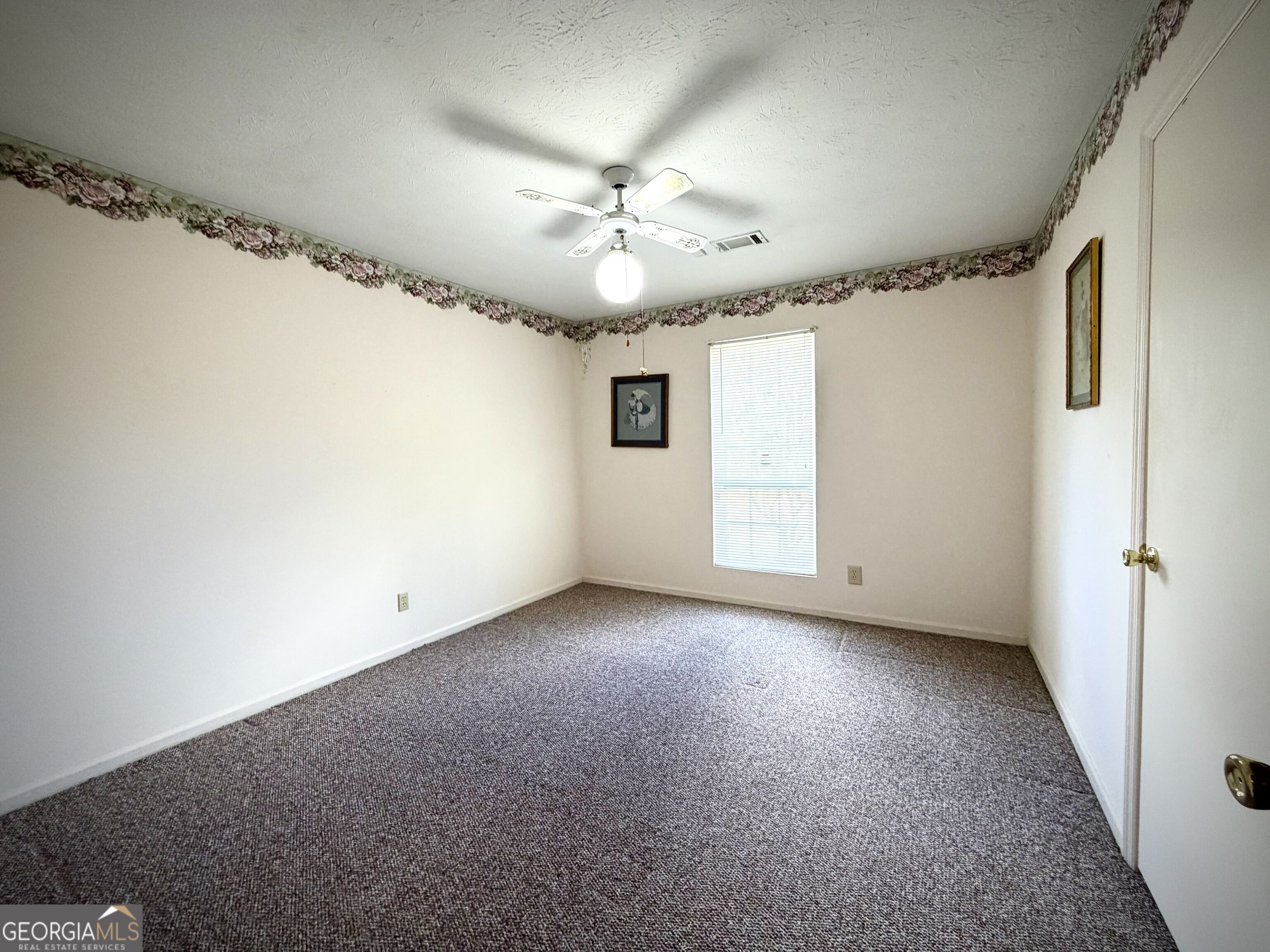 Empty carpeted room with a window, ceiling fan, and floral wallpaper border.