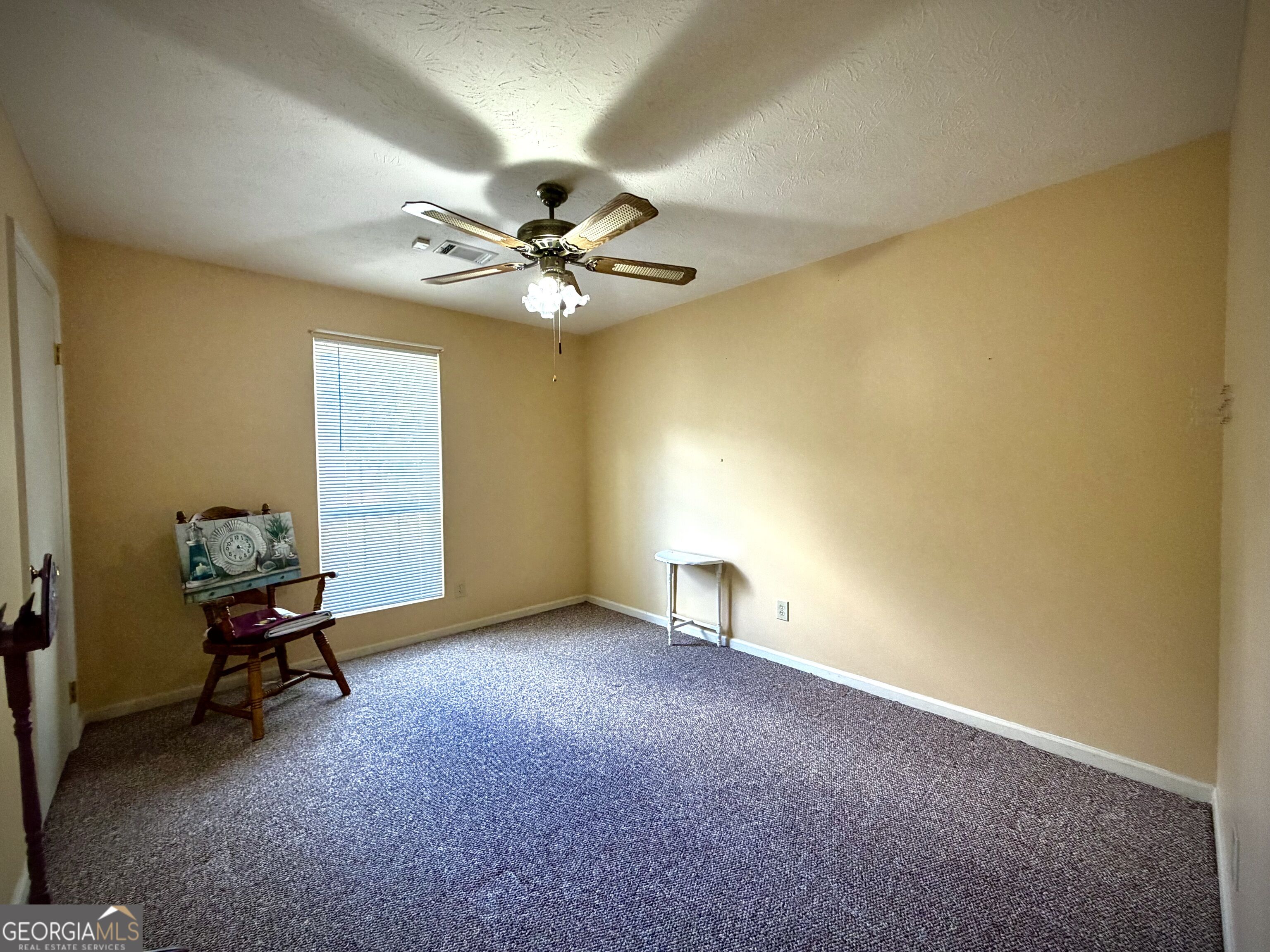 Mostly empty bedroom with gray carpet, beige walls, a ceiling fan, and a window with blinds.