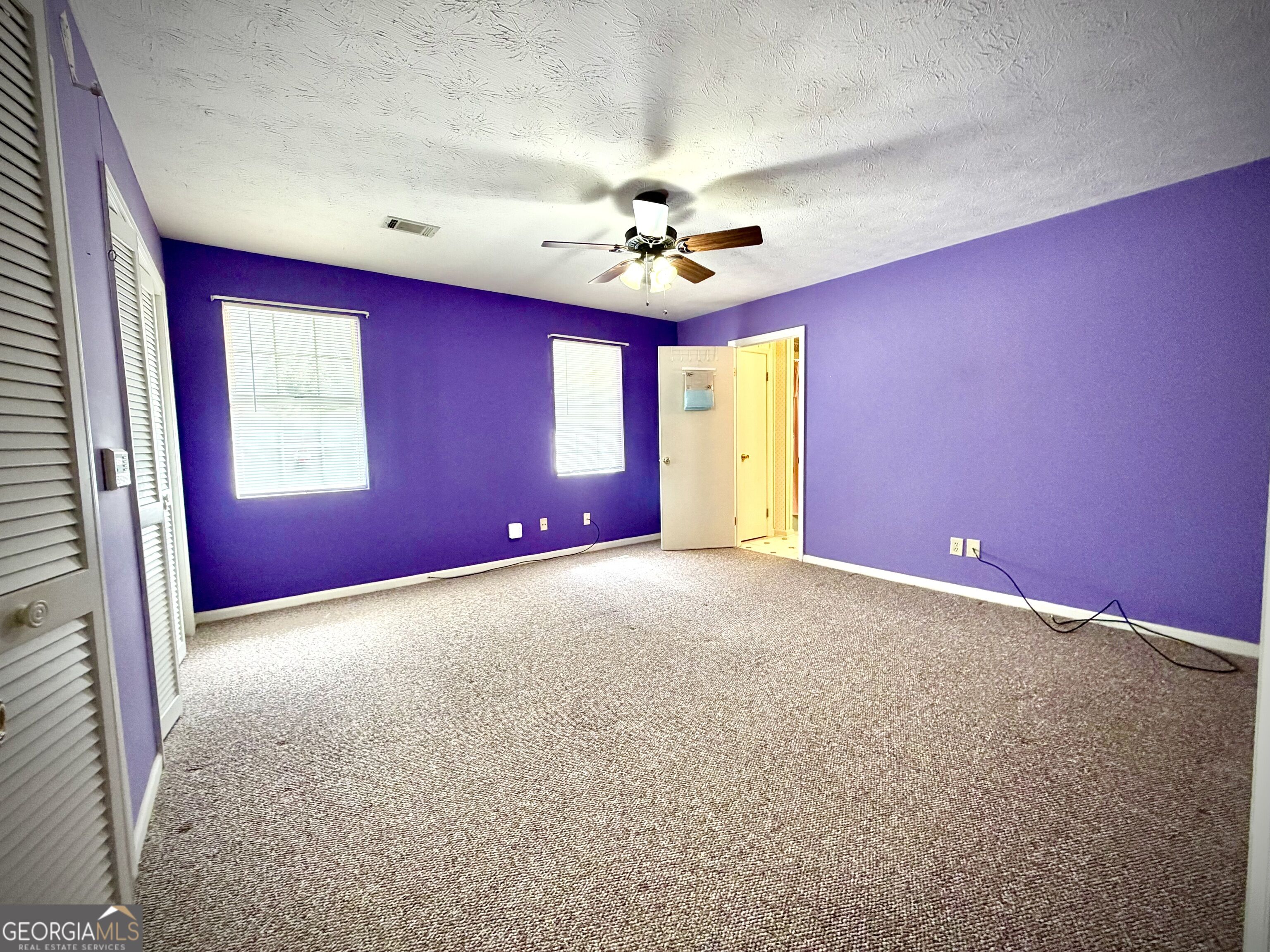 Empty bedroom with purple walls, beige carpeting, two windows, and a ceiling fan.