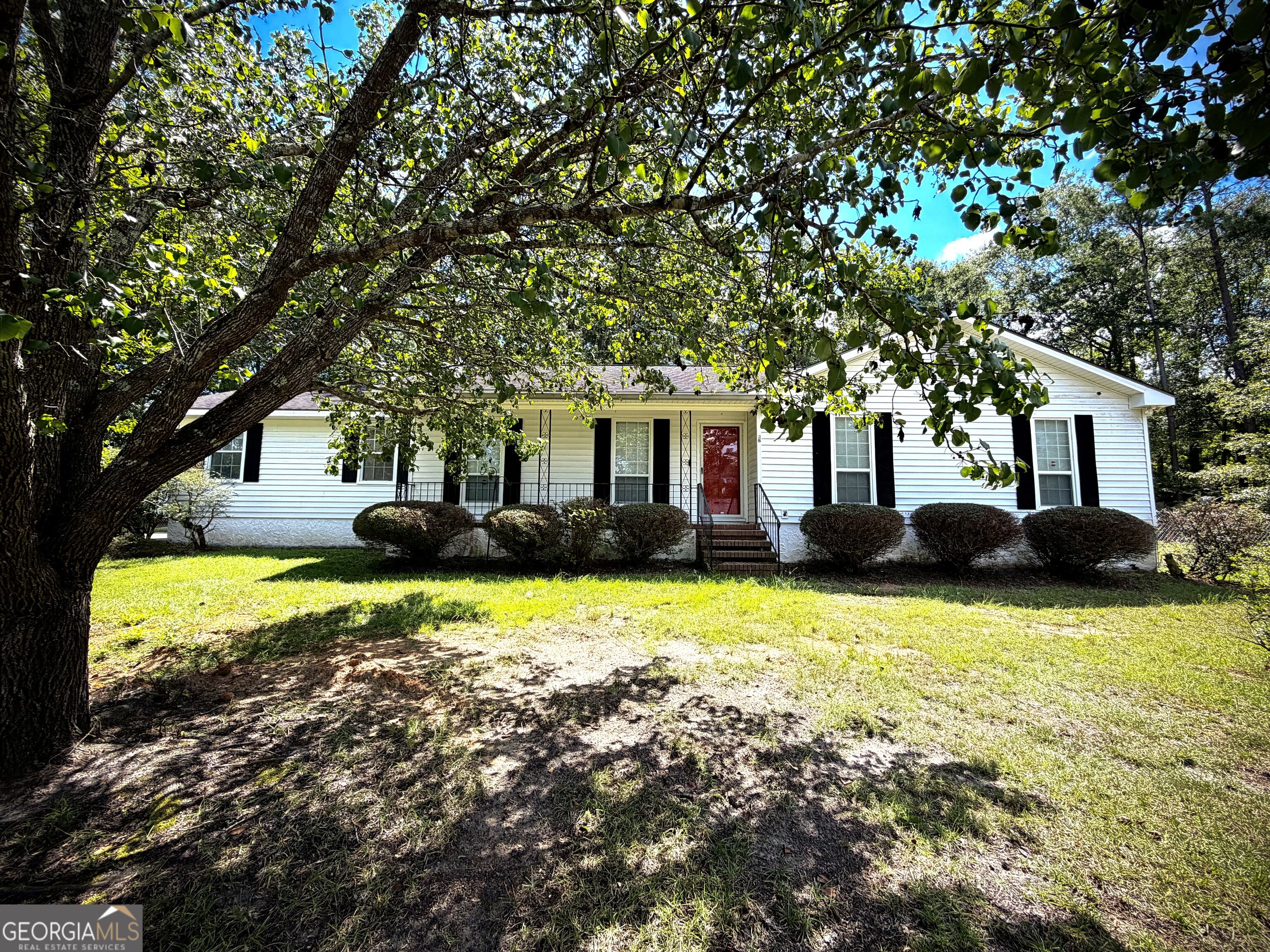 White single-story home with a red front door, covered porch, and large shade tree.