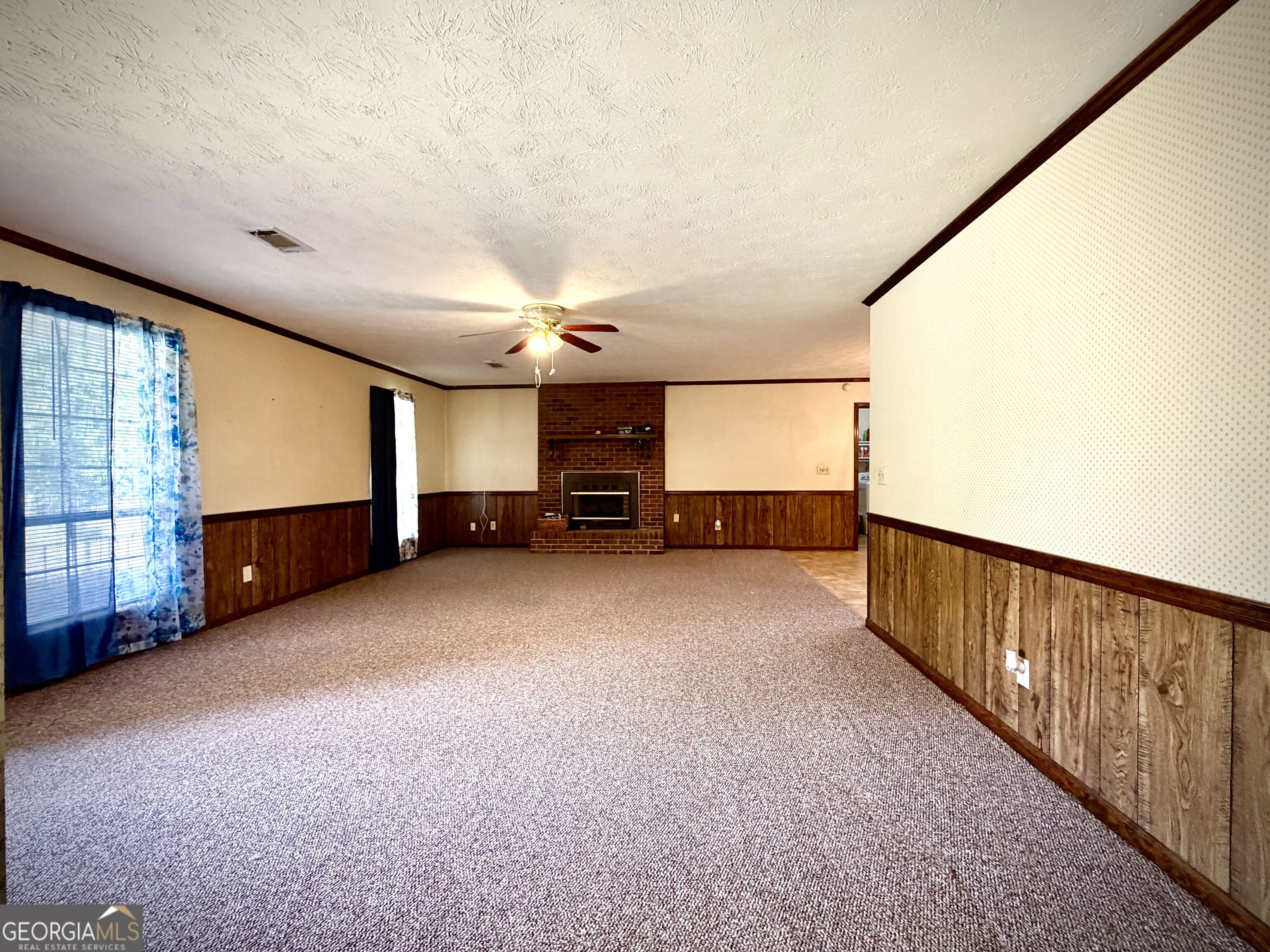 Empty living room with a brick fireplace, carpet, wood-paneled walls, and ceiling fan.