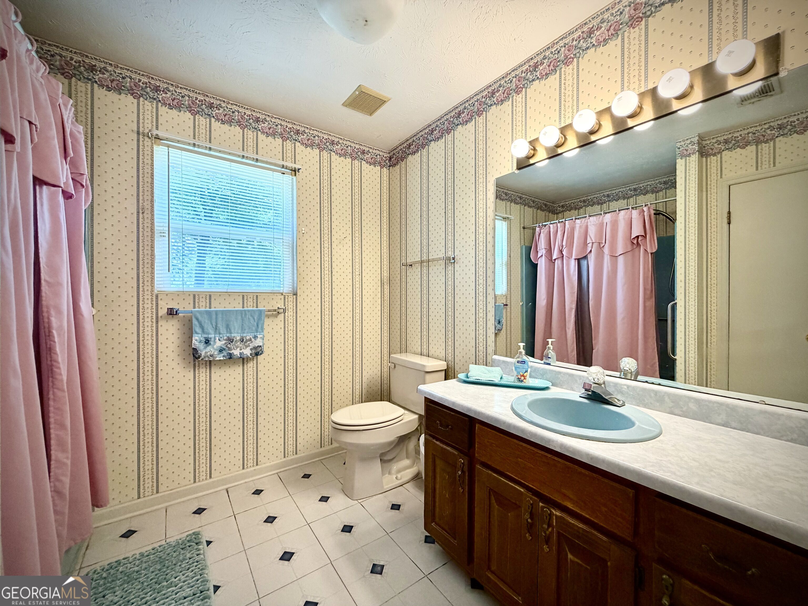 Bathroom with a wood vanity and blue sink, pink shower curtain, and patterned wallpaper.