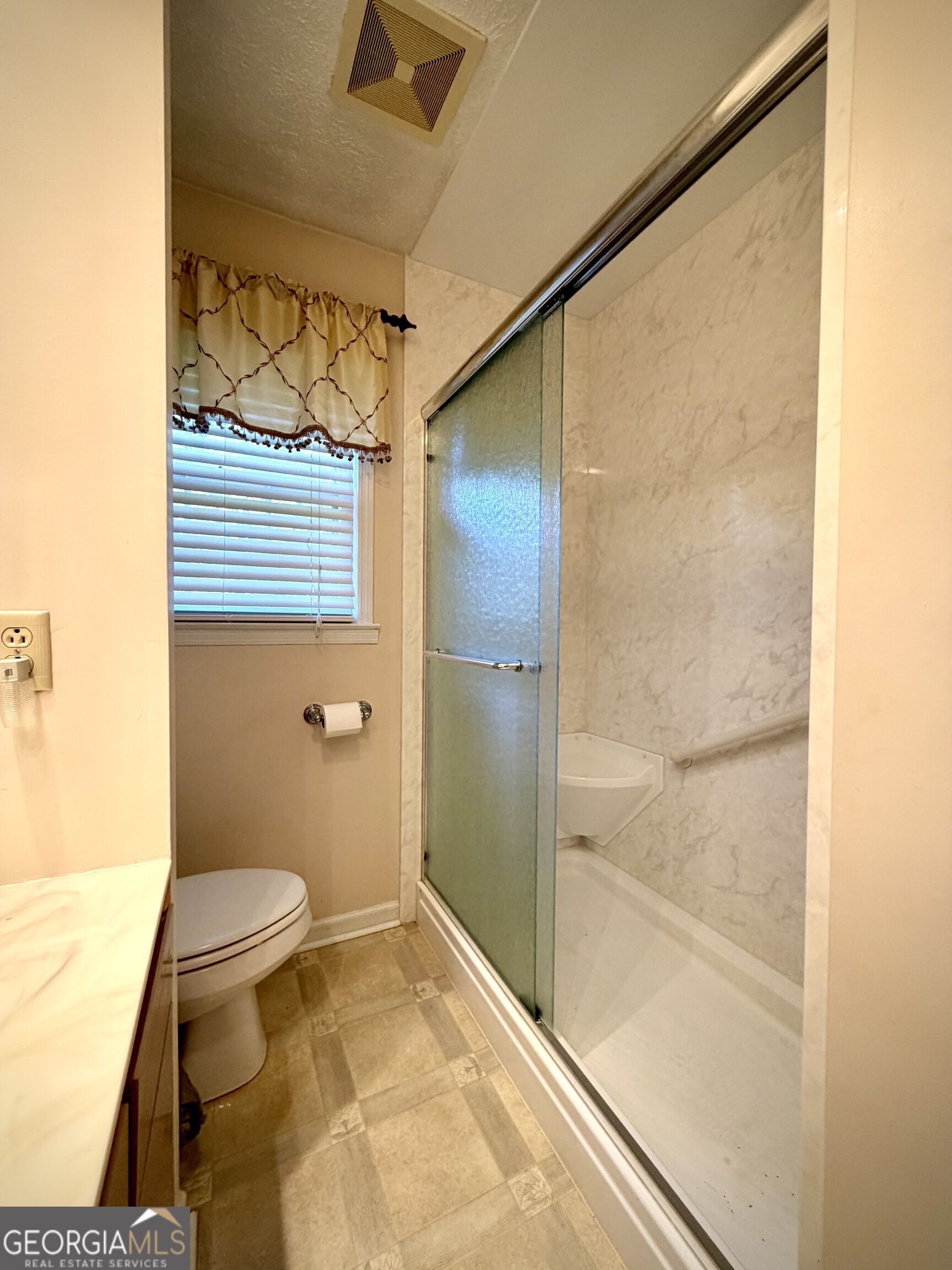 Bathroom with a glass shower enclosure, marble-patterned walls, grab bar, and window with blinds.