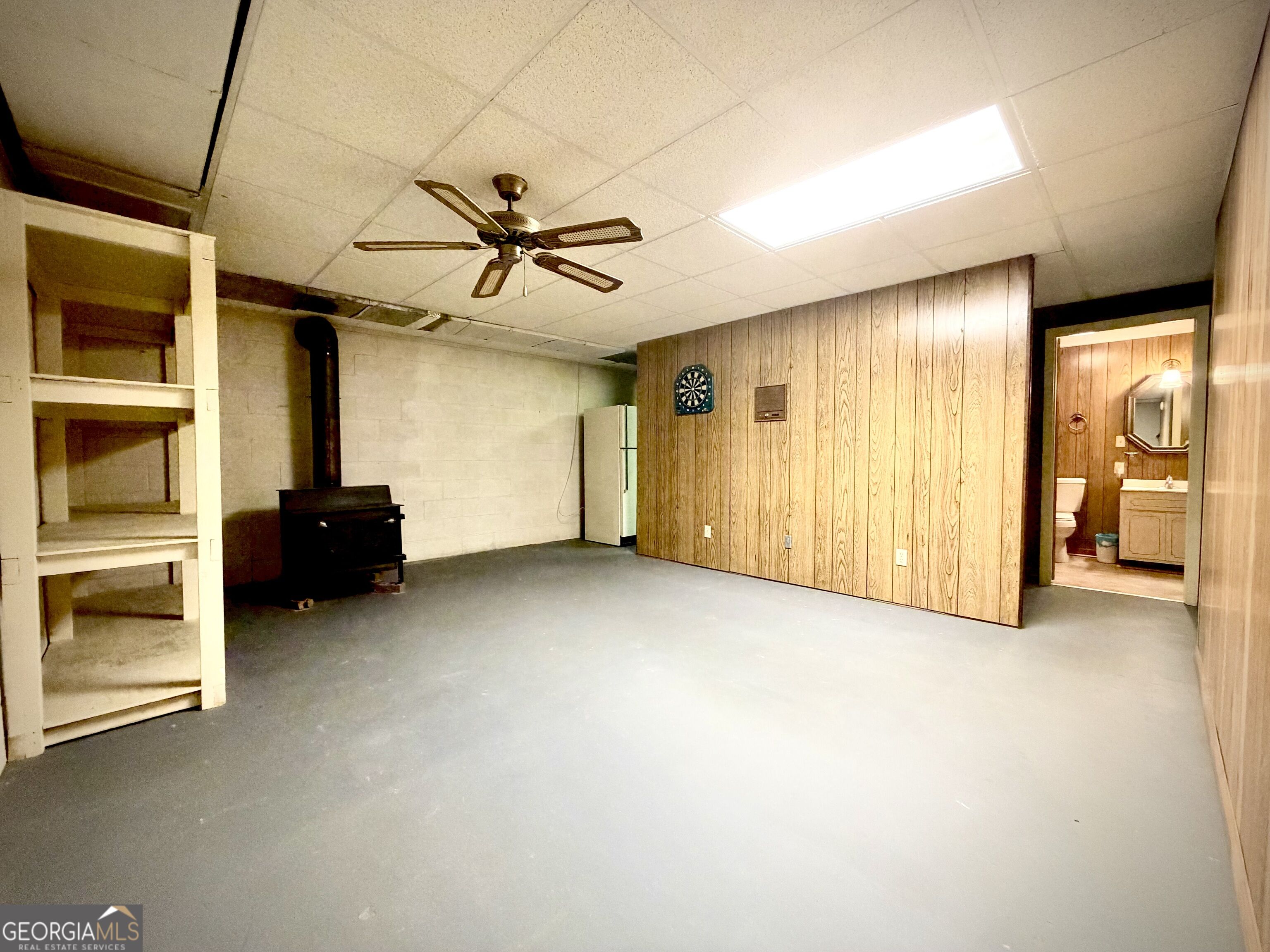 Basement recreation room with wood-paneled walls, gray flooring, a wood-burning stove, and an adjoining bathroom.