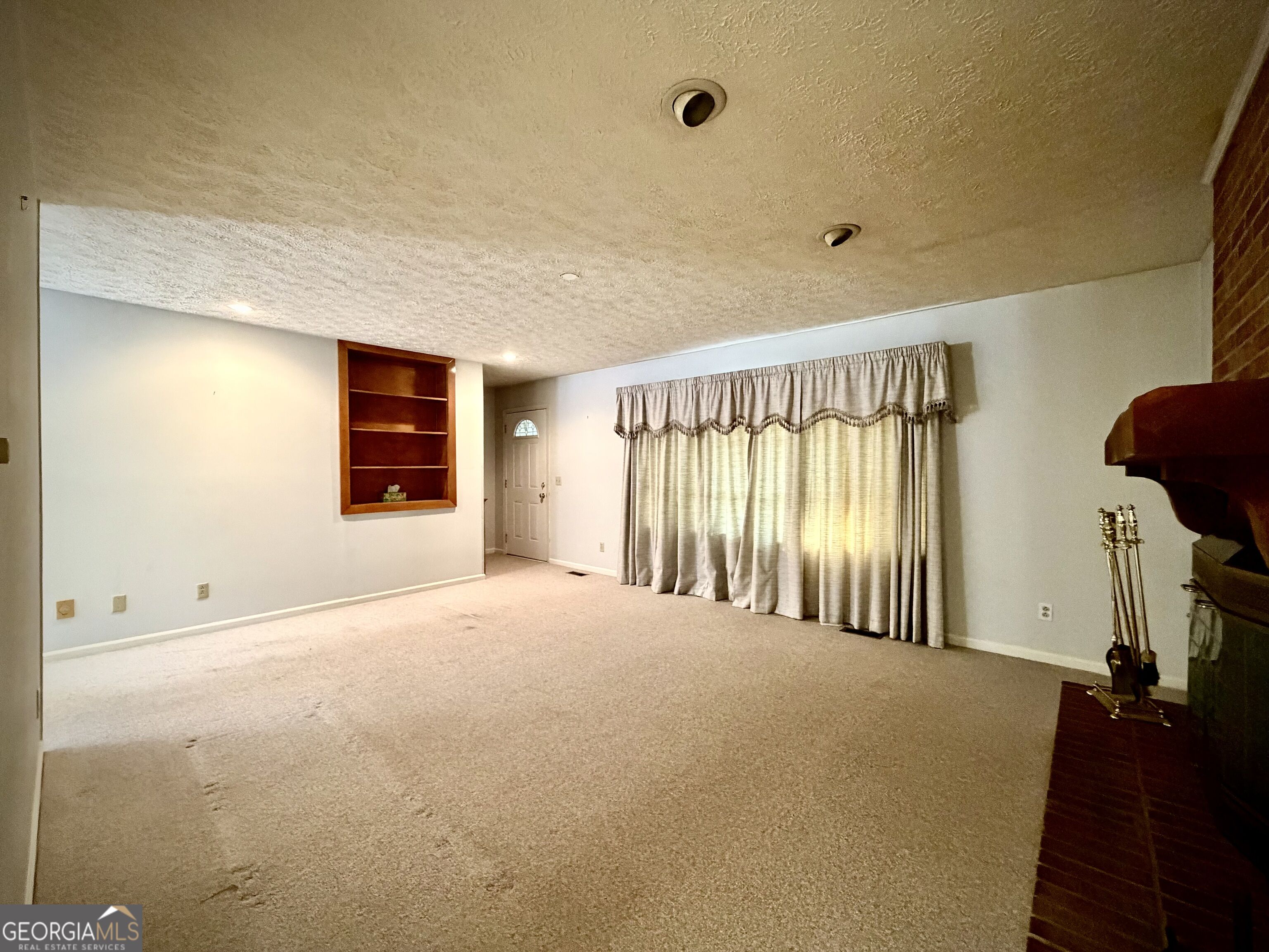 Empty carpeted living room with a built-in wooden bookshelf, curtained windows, and brick fireplace.