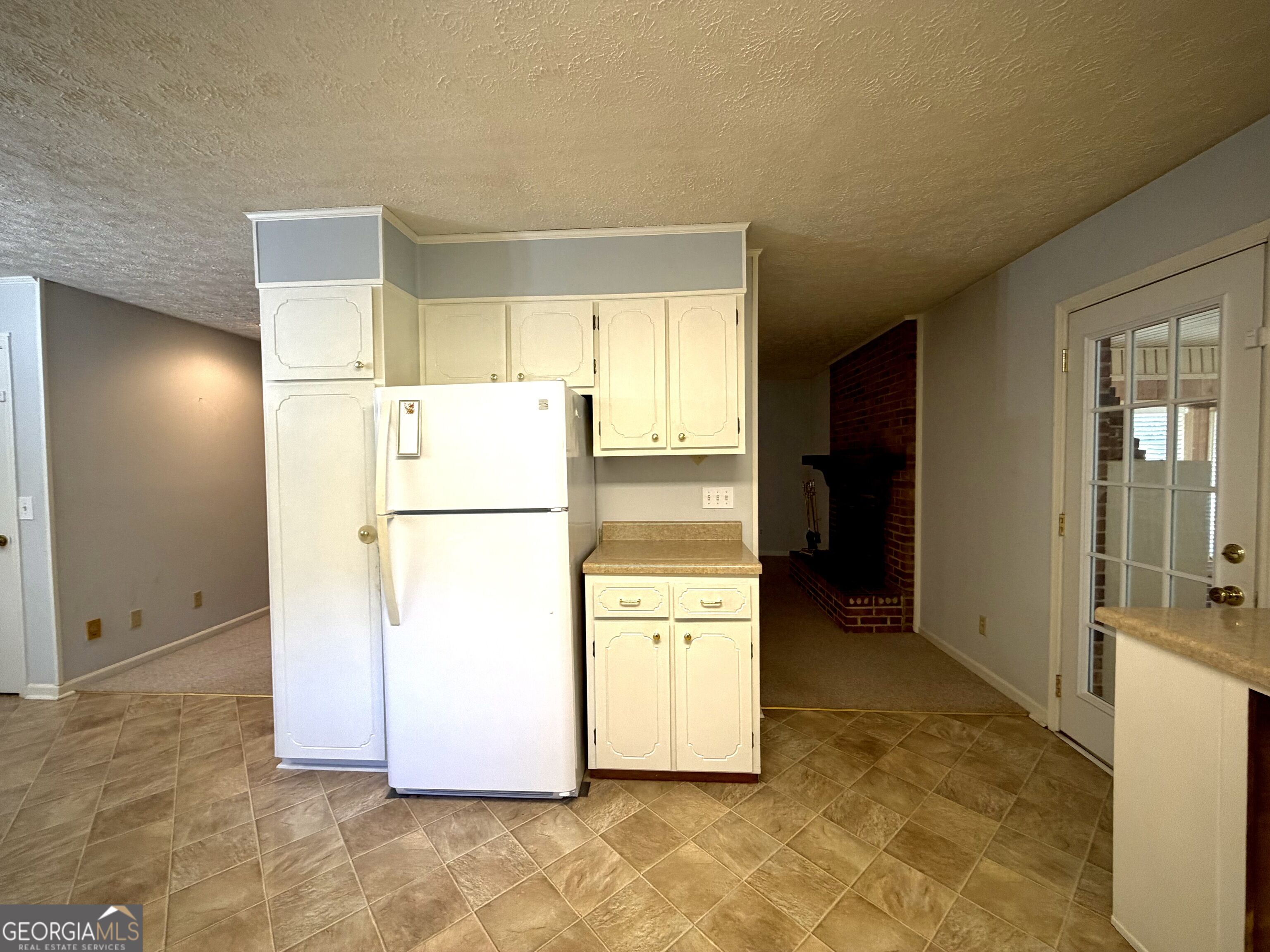 Kitchen with white cabinets and refrigerator, tan tile flooring, and a brick fireplace nearby.