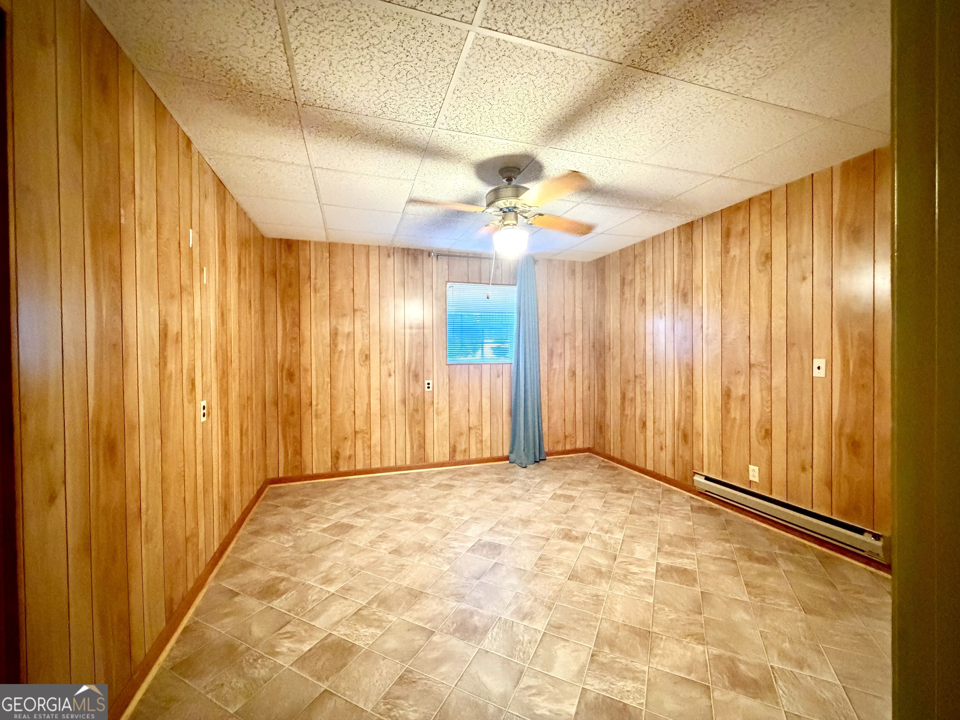 Empty wood-paneled room with tiled flooring, a ceiling fan, and a small curtained window.