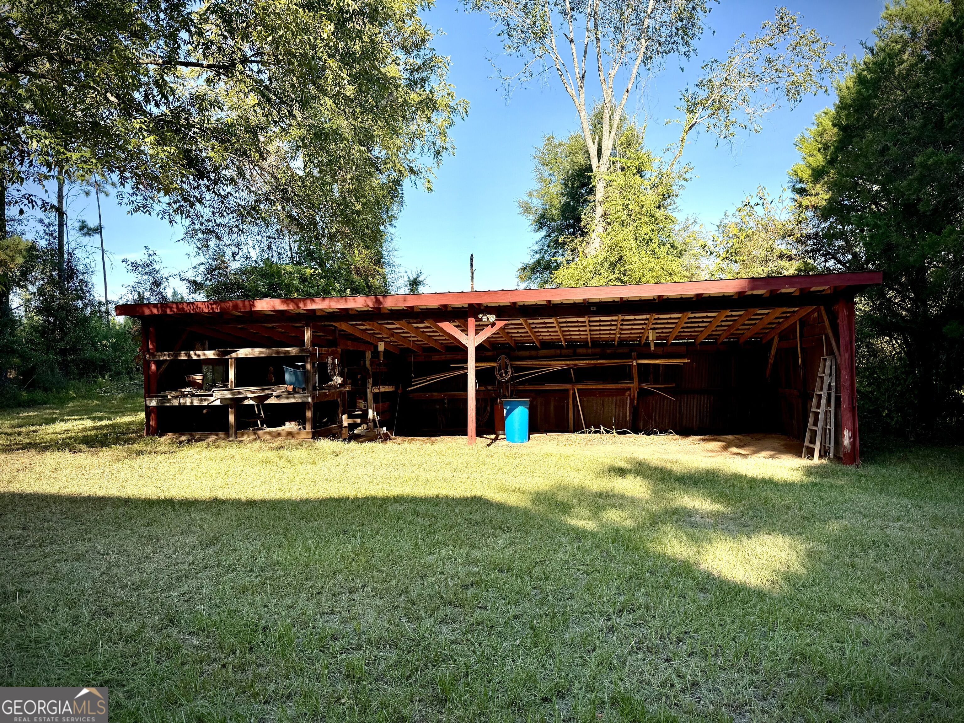 Open-sided red timber barn with a broad roof, tools, and a blue barrel in a grassy yard.