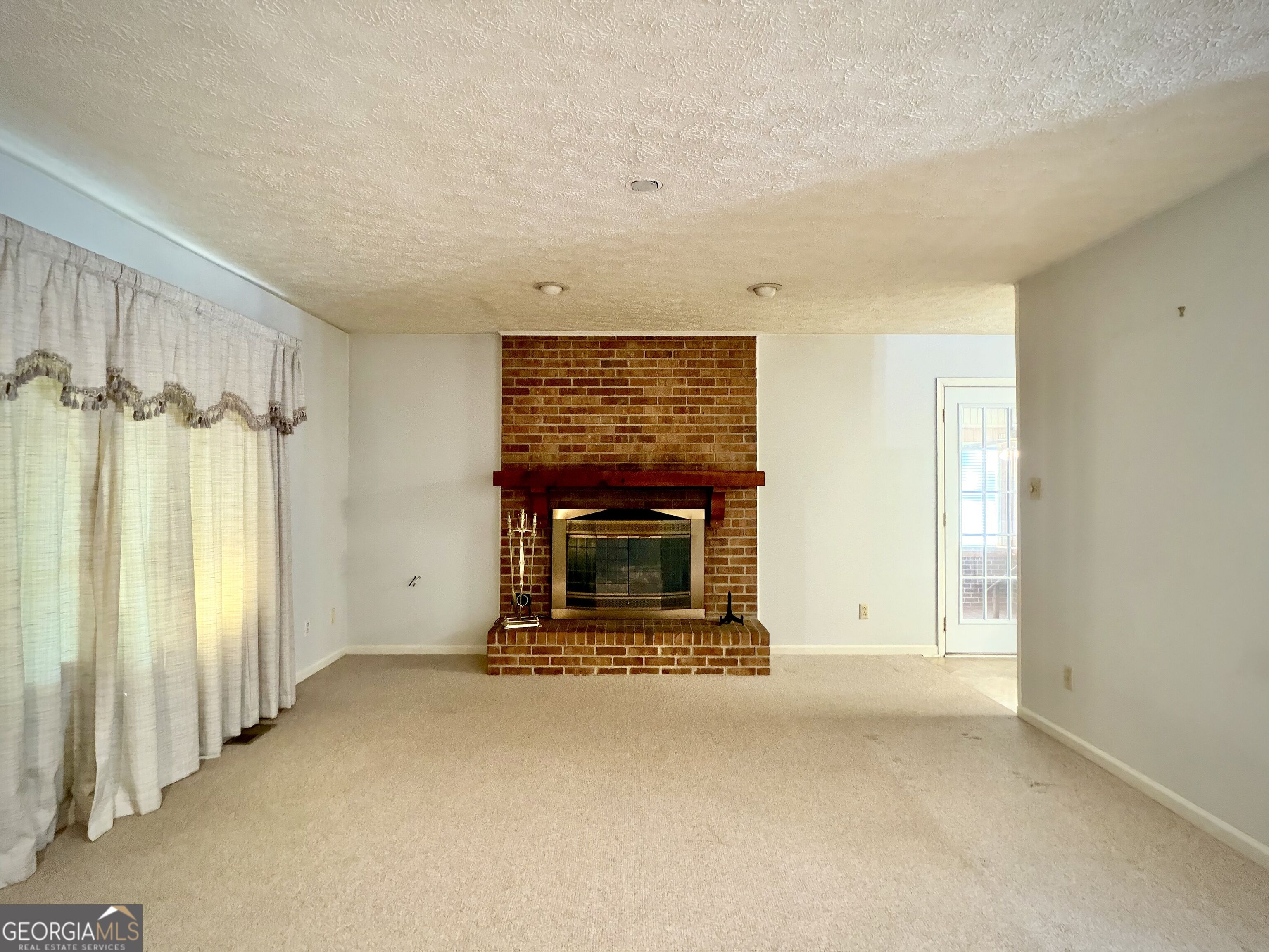 Empty living room with a red brick fireplace, dark wood mantel, and beige carpet.