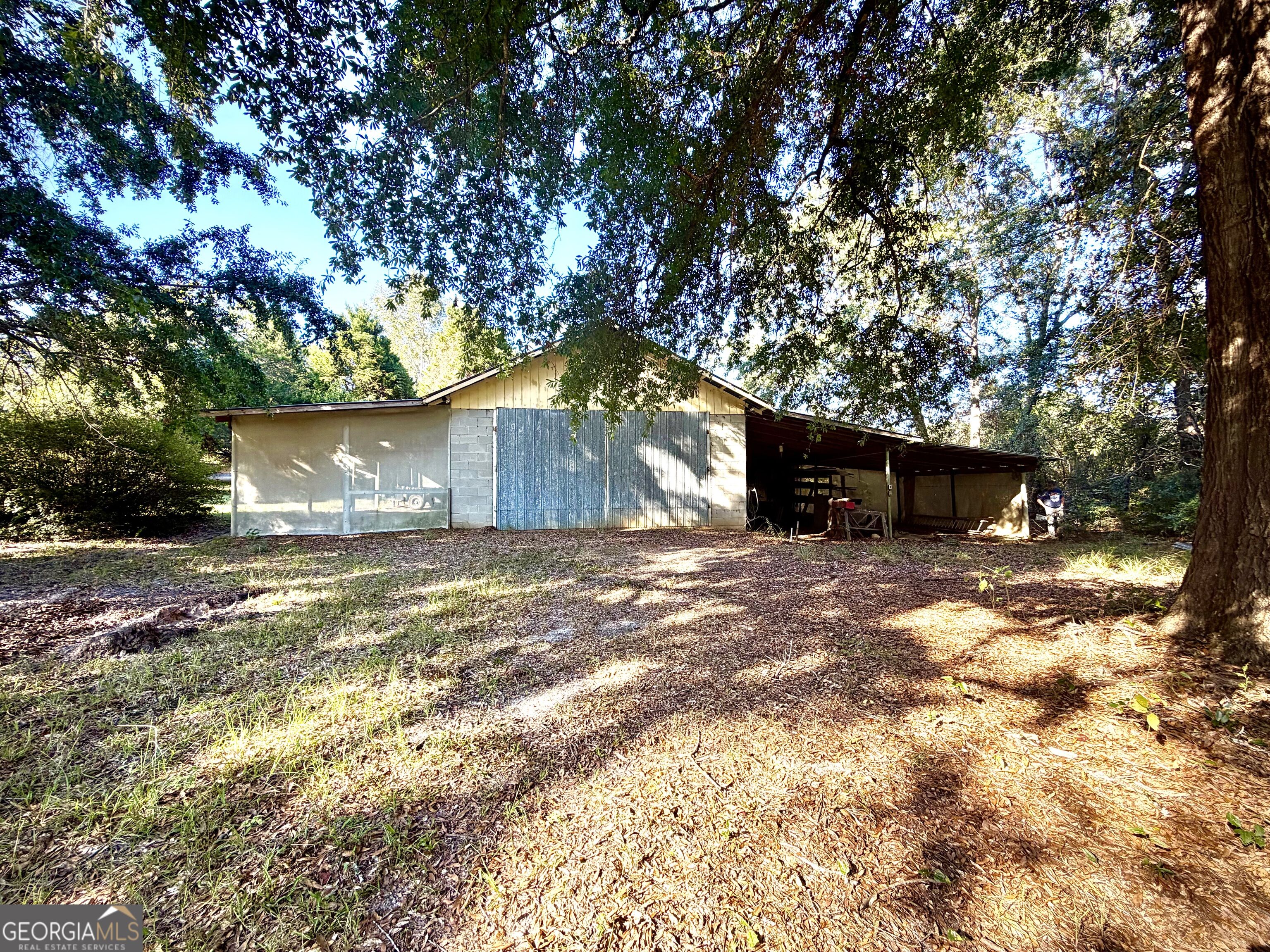 Detached utility building with large metal doors and an open-sided covered area beneath trees.