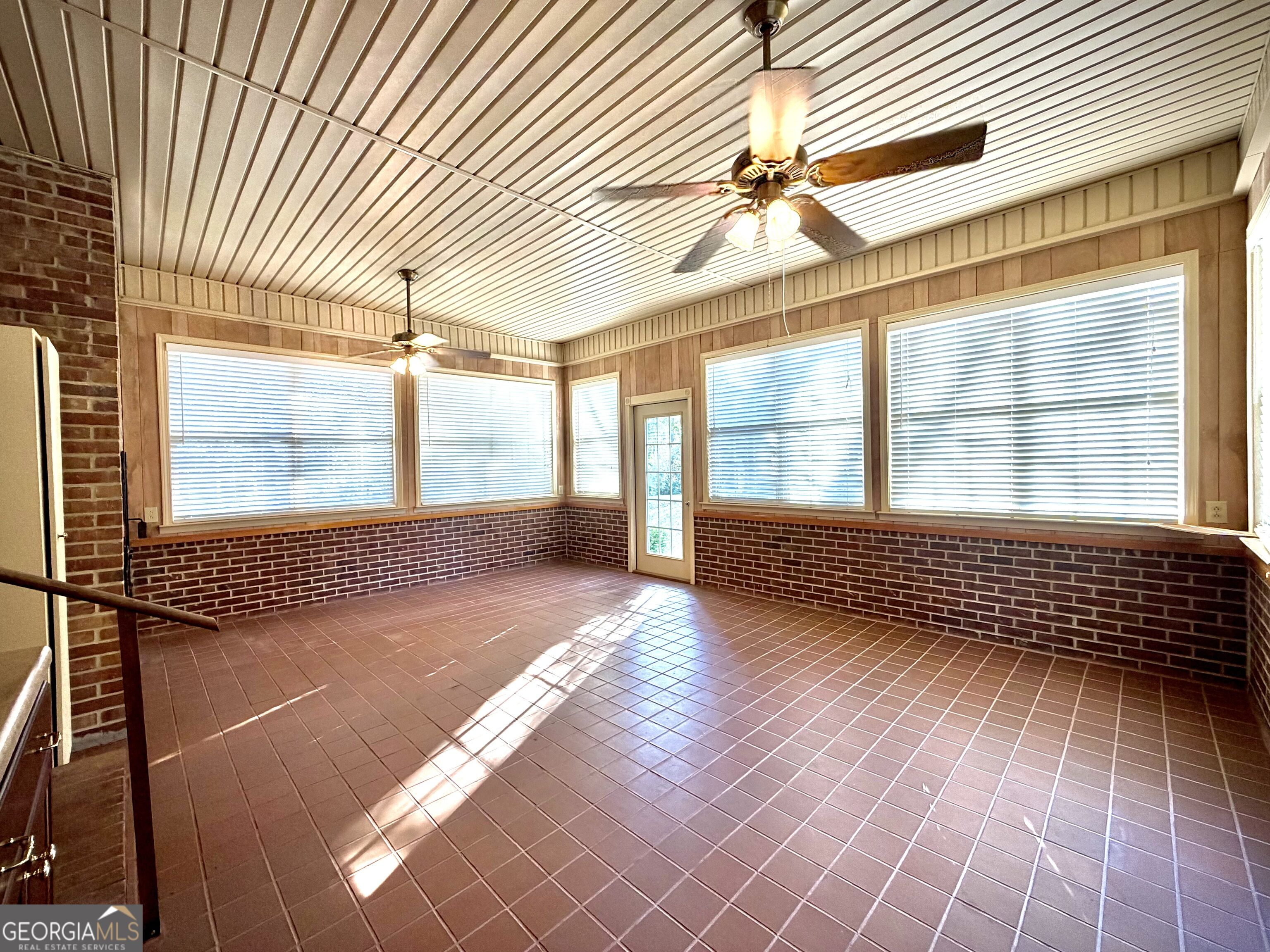 Empty sunroom with brick wainscoting, tiled floor, broad windows, and two ceiling fans.