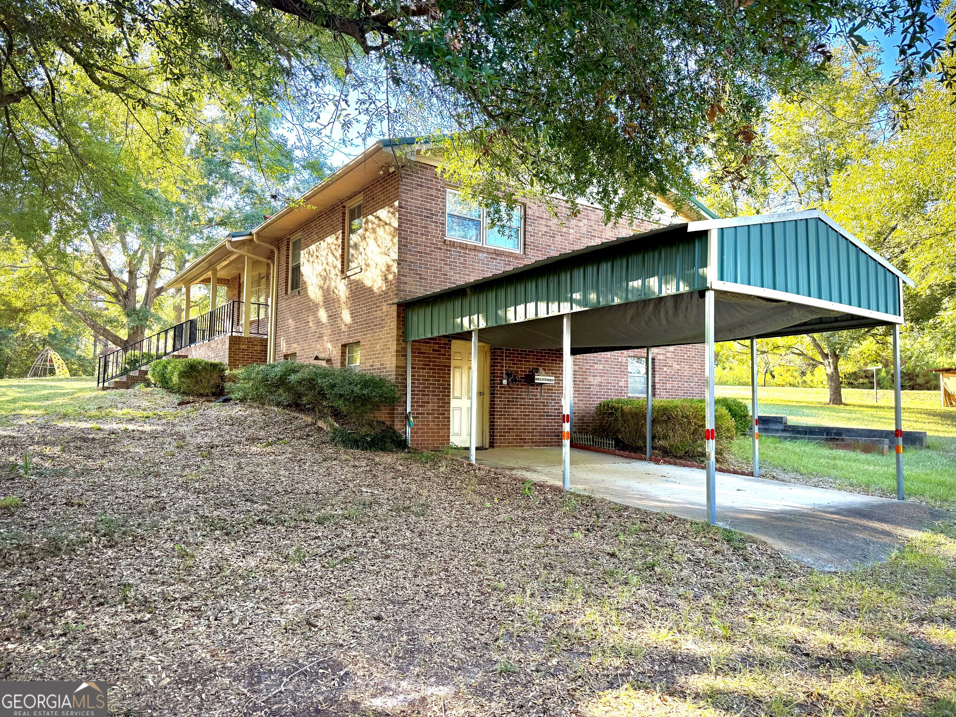 Two-story brick house with a covered porch and large green metal carport beneath mature trees.