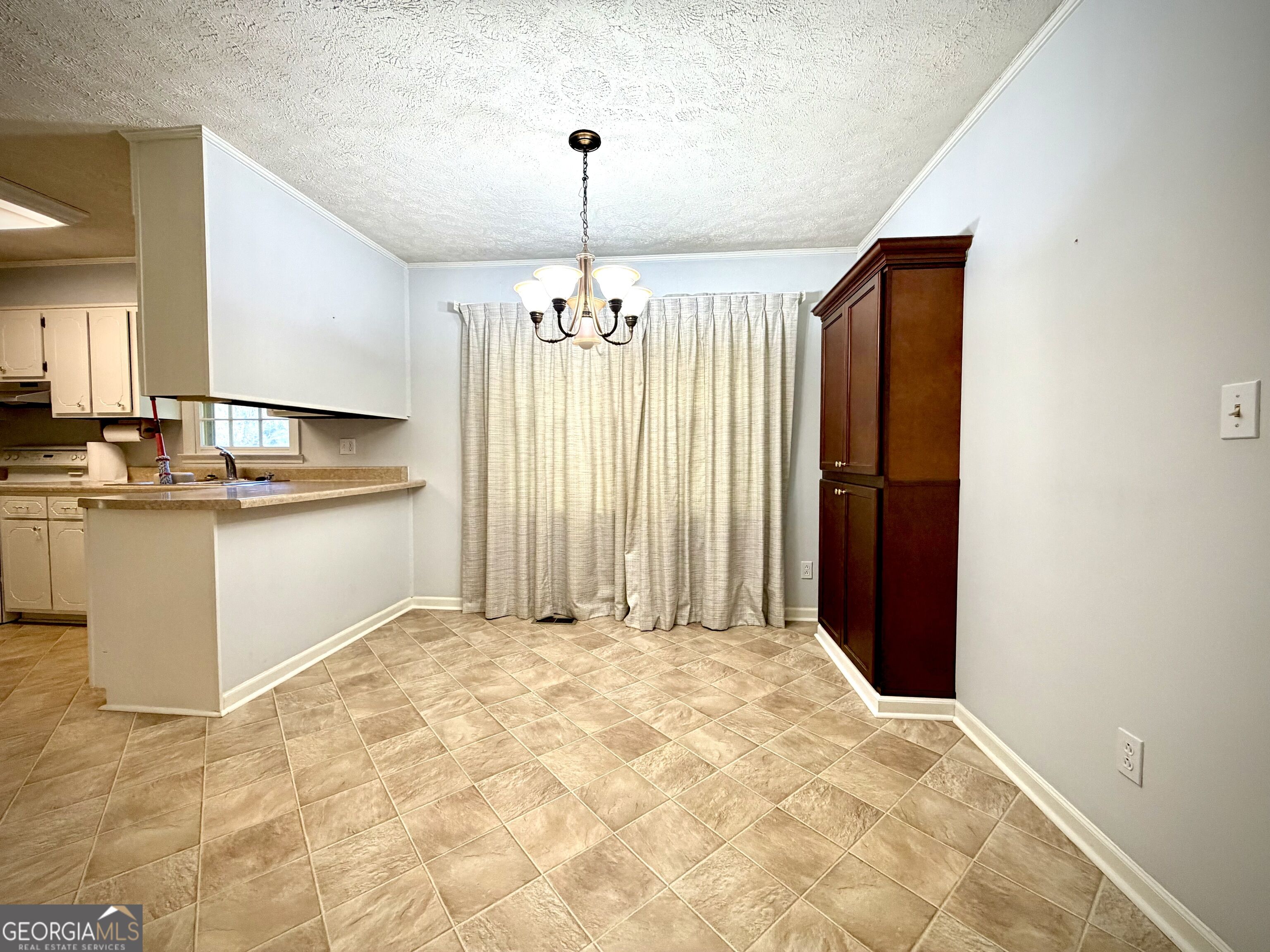 Dining area with tan tile floors, a chandelier, curtained window, and tall dark wood cabinet.