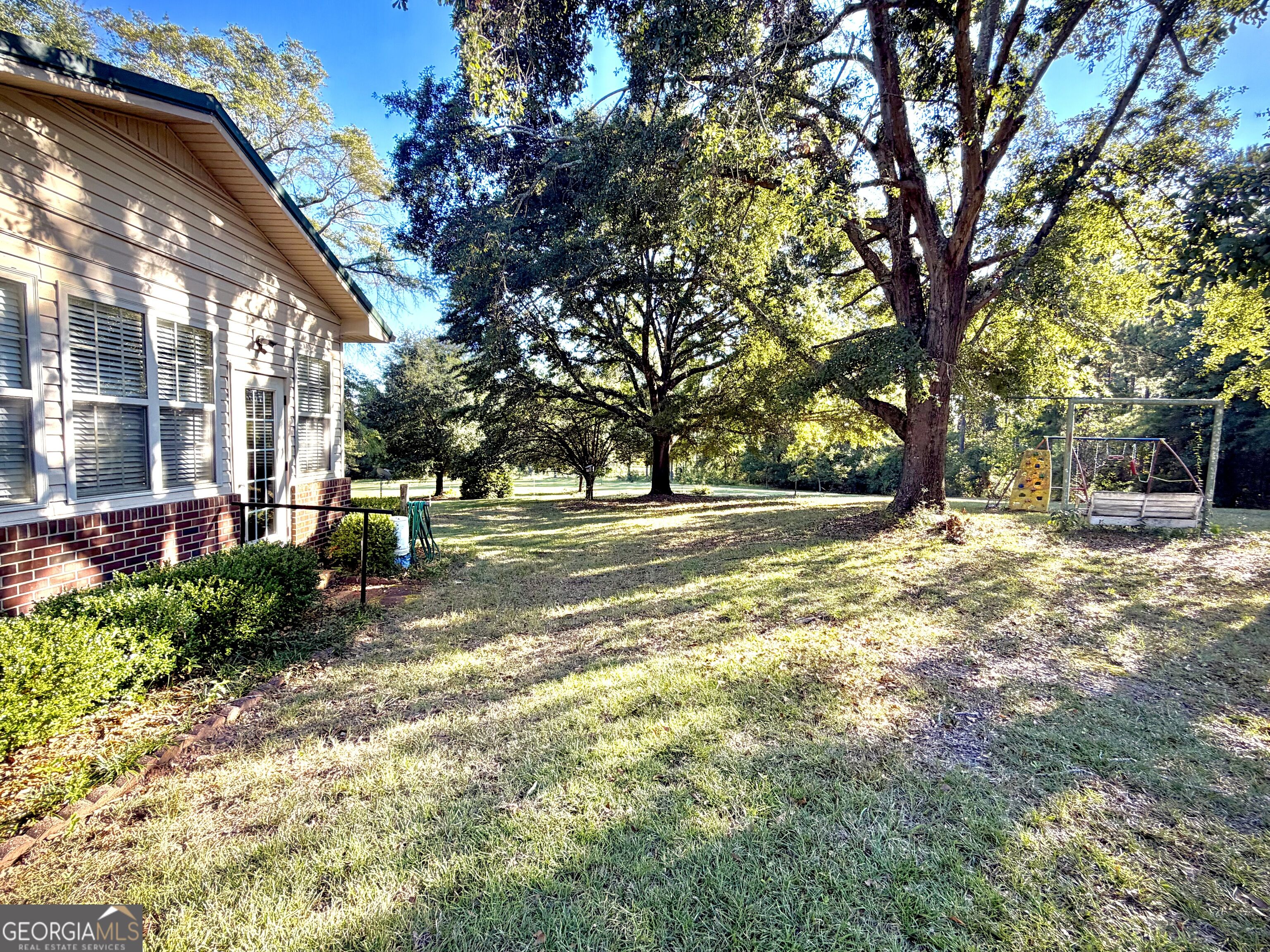 Grassy backyard beside a light-sided home, shaded by mature trees with a children's playset.