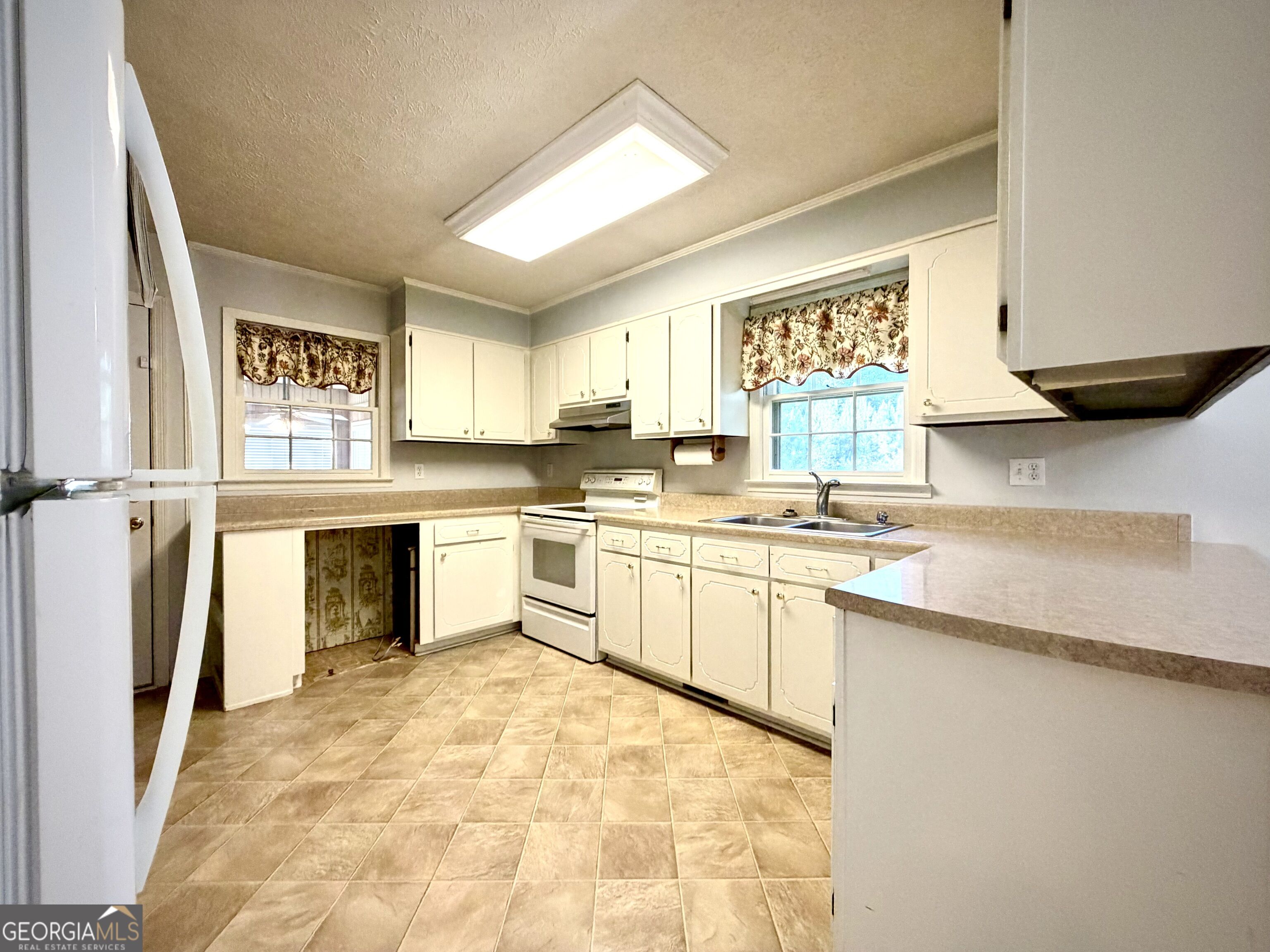 Vacant kitchen with white cabinets, tan tile floors, a white stove, and a sink beneath a window.