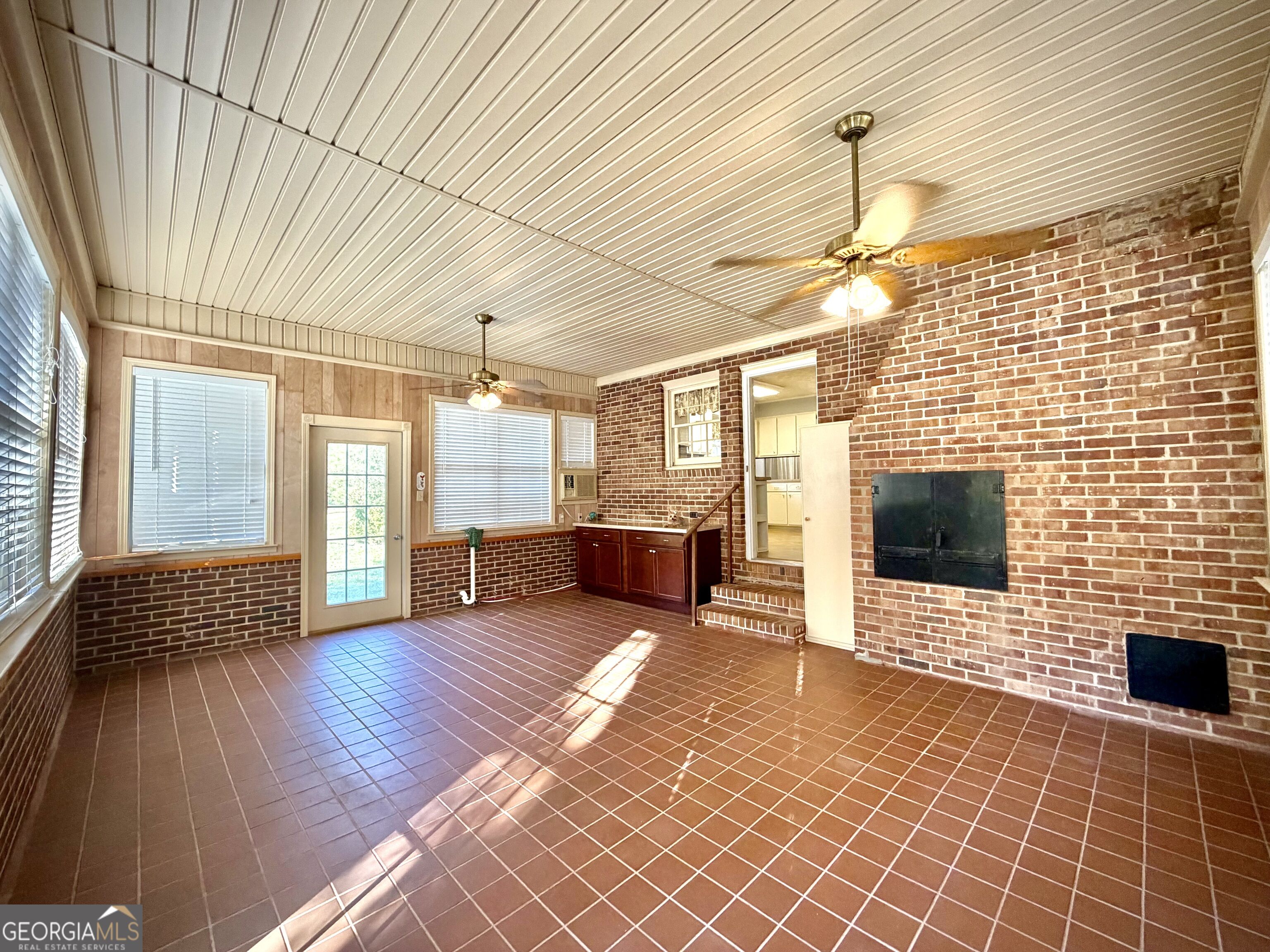 Vacant sunroom with a brick fireplace, tiled floor, ceiling fans, and a small cabinet counter.