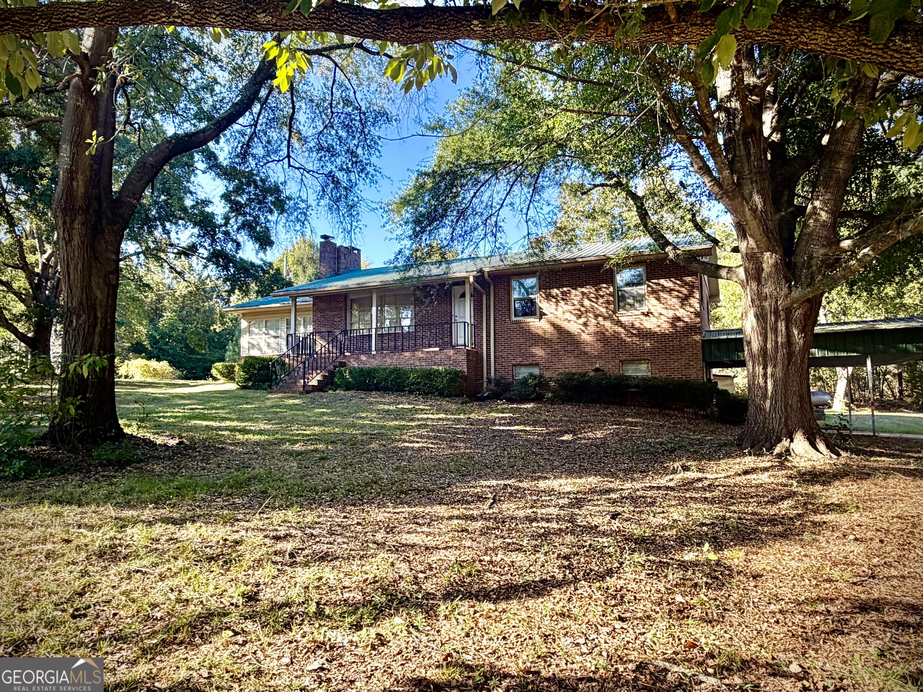 Red brick house with a covered porch and carport beneath large shade trees.