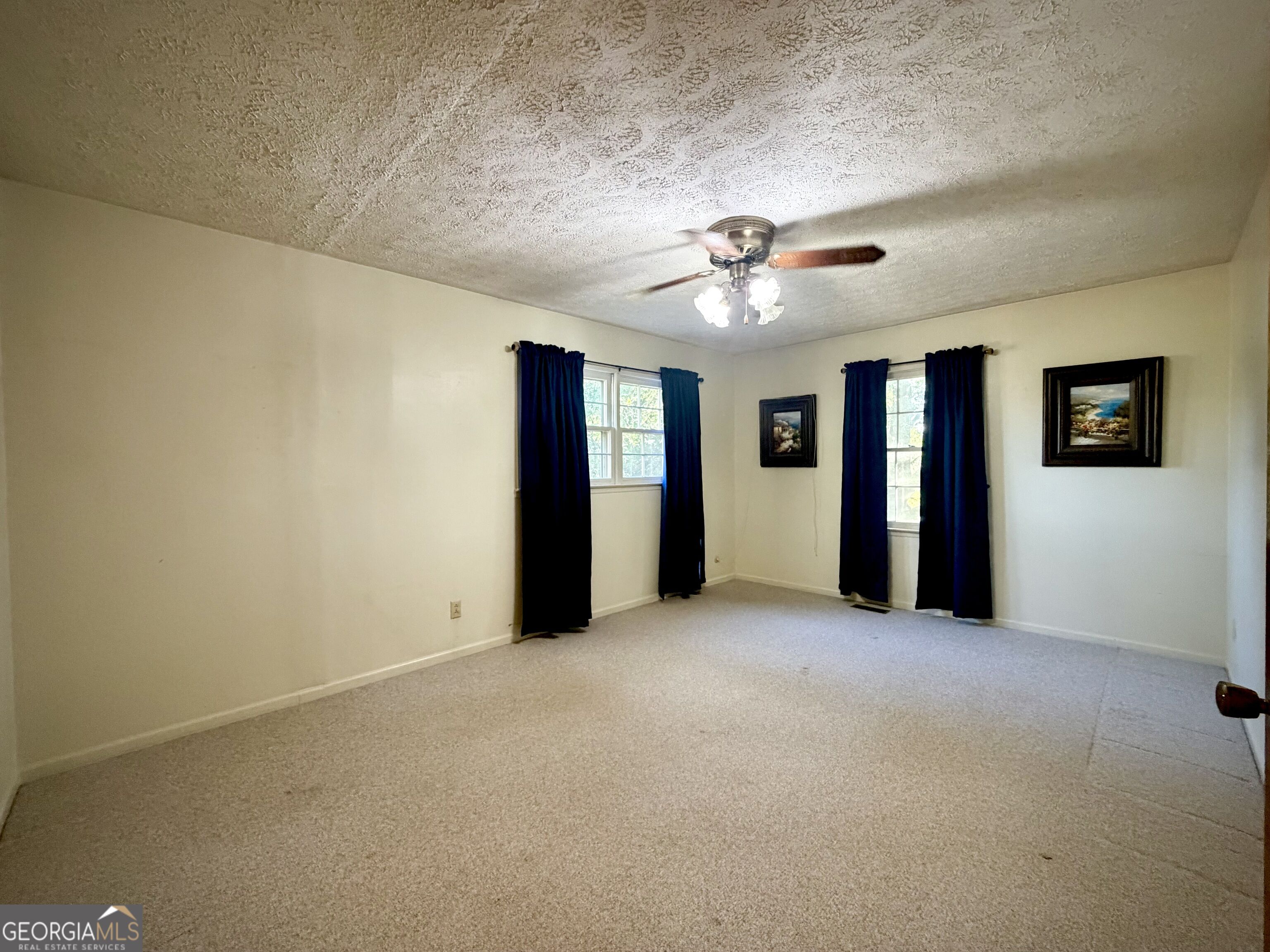 Empty carpeted room with blue curtains, two windows, and a ceiling fan with lights.