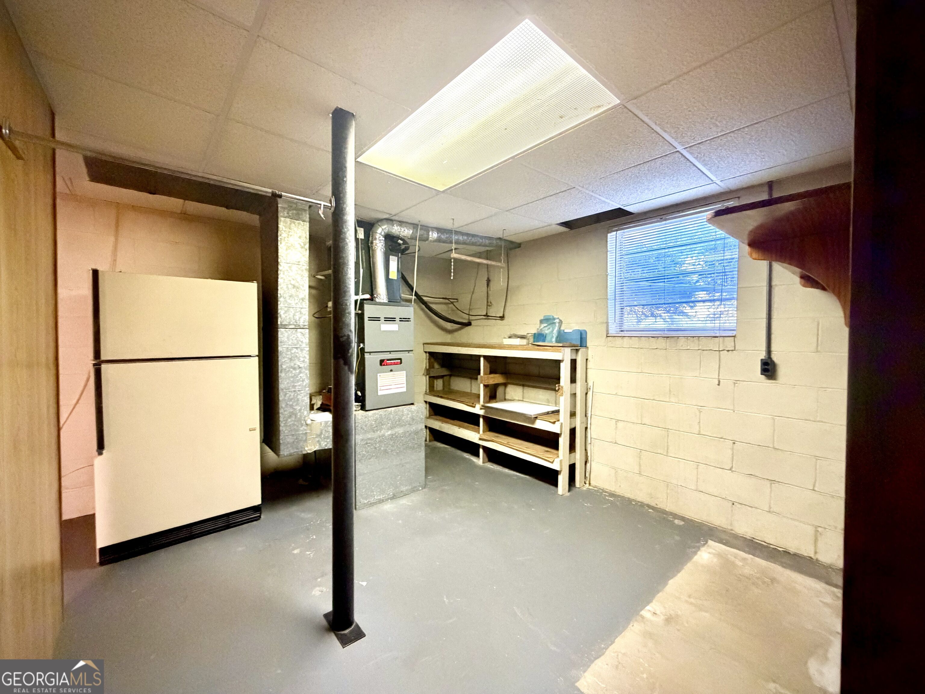Basement utility room with a refrigerator, furnace, wooden storage shelves, and a central support post.