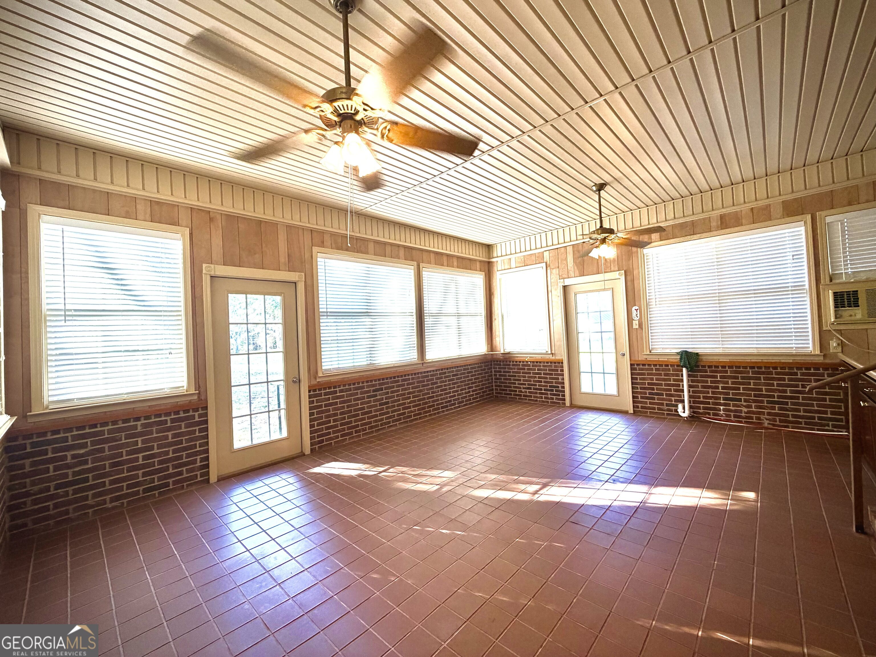 Empty sunroom with a reddish tile floor, brick lower walls, numerous windows, and two ceiling fans.