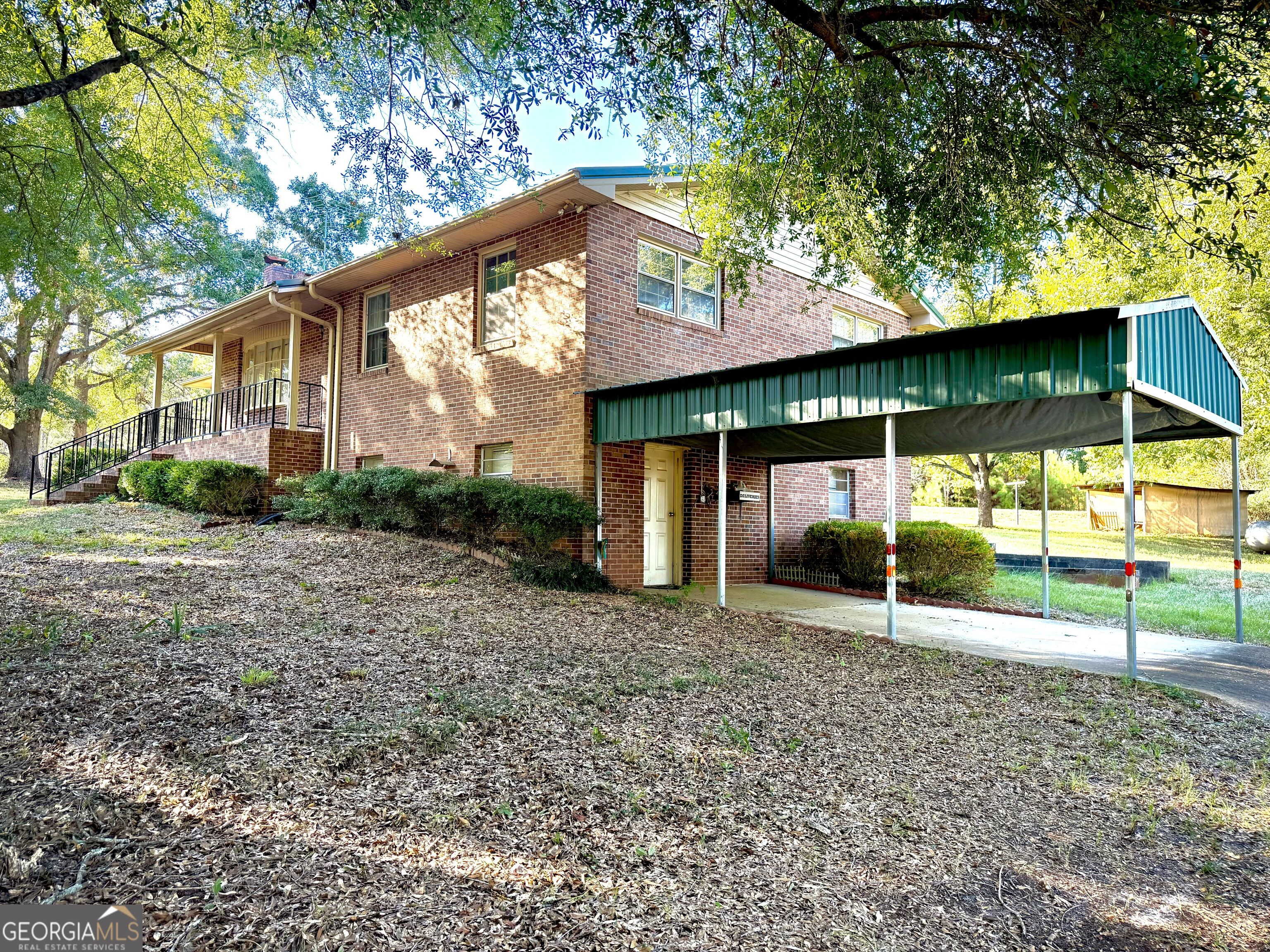 Two-story brick house with a covered porch and large green metal carport beneath mature trees.