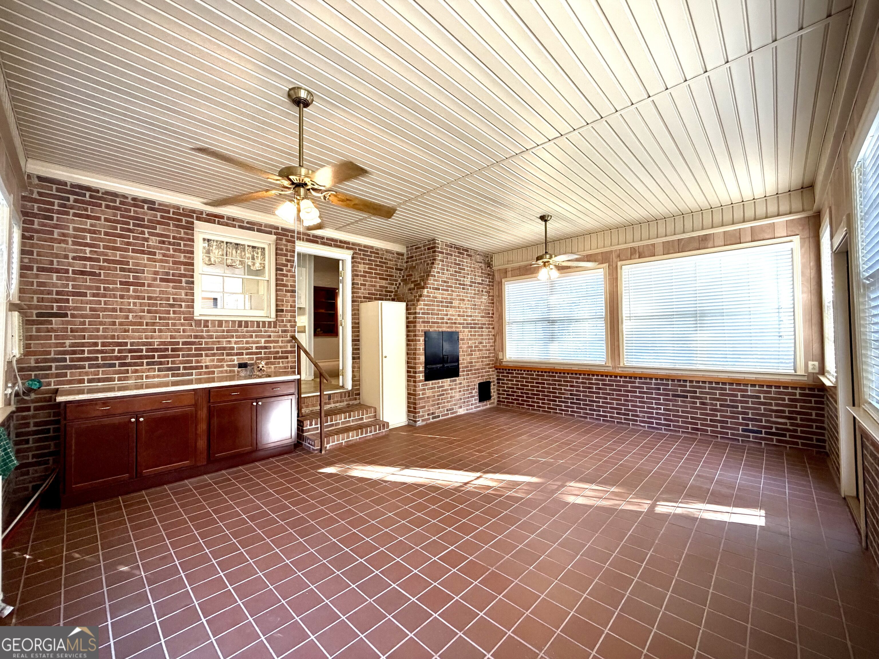 Enclosed brick sunroom with tiled floors, large windows, ceiling fans, and dark wood cabinets.