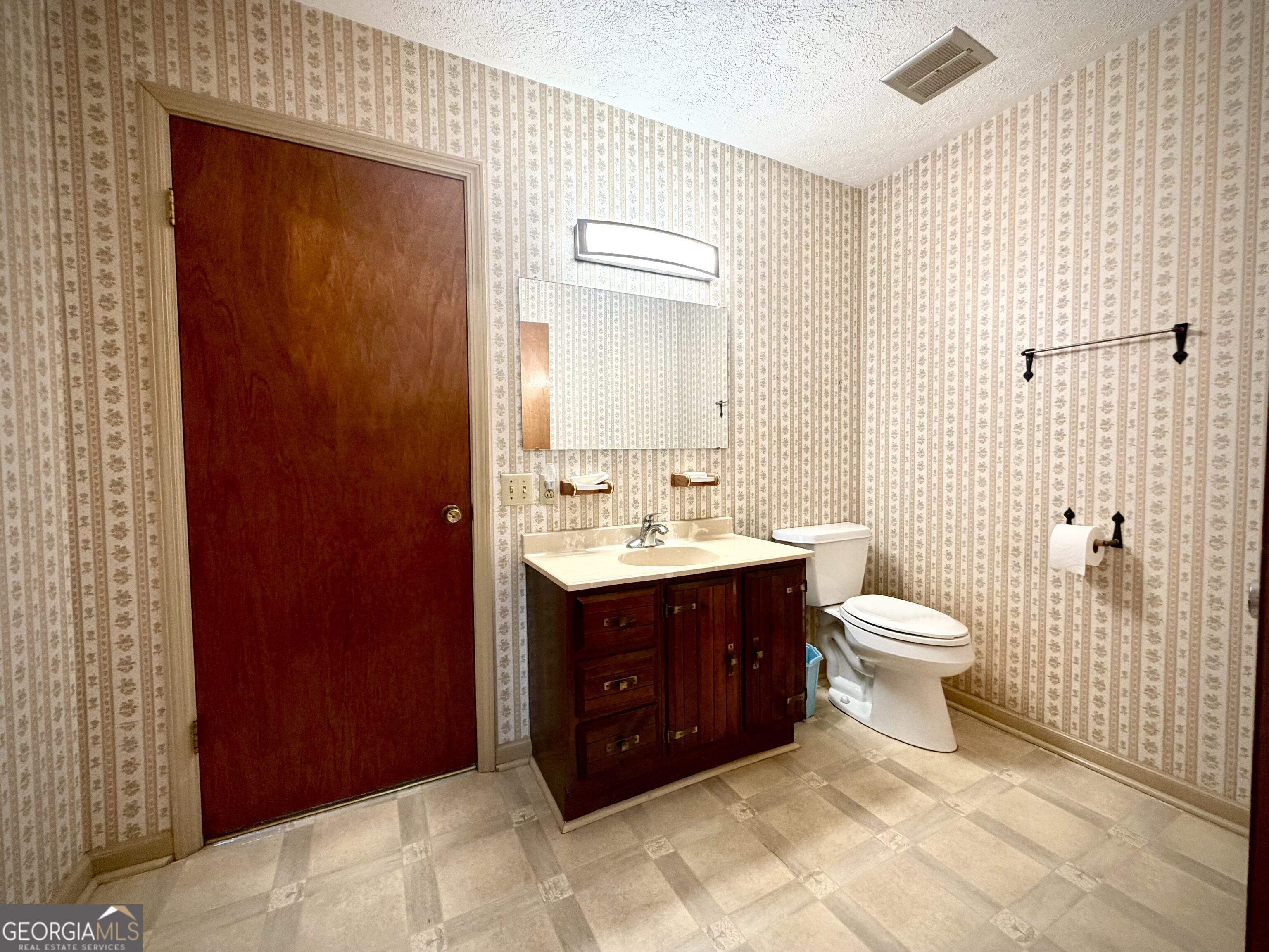 Bathroom with patterned wallpaper, dark wood vanity, white toilet, and beige tiled floor.
