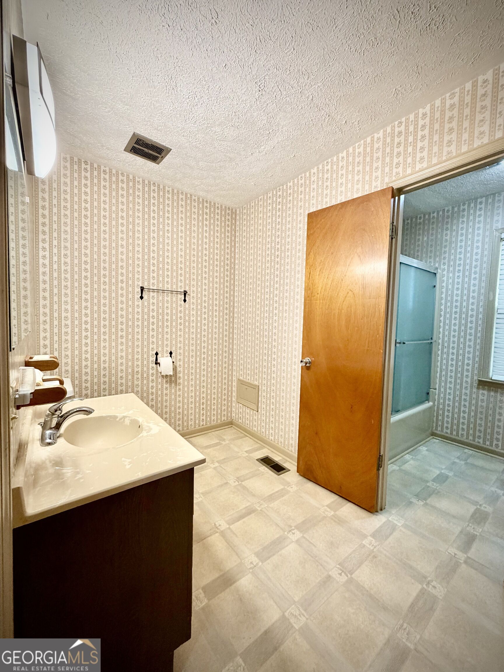 Bathroom with a single-sink vanity, patterned wallpaper, and a tub-shower behind the open door.