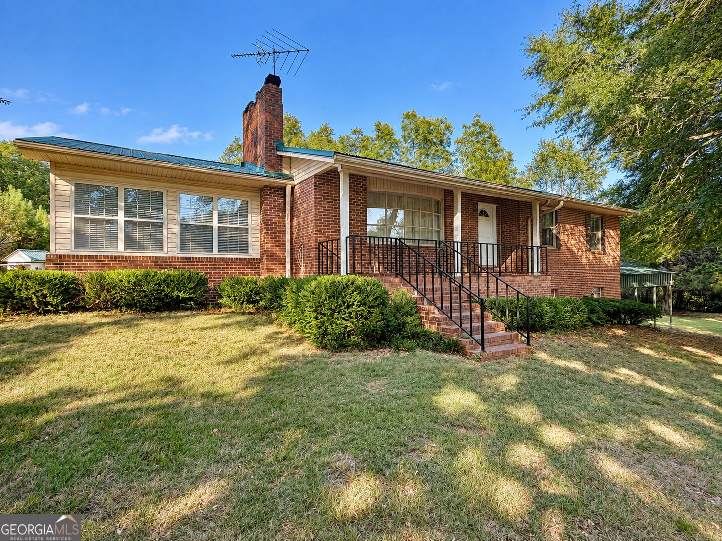 Brick ranch home with a raised porch, black railings, front lawn, and mature trees.