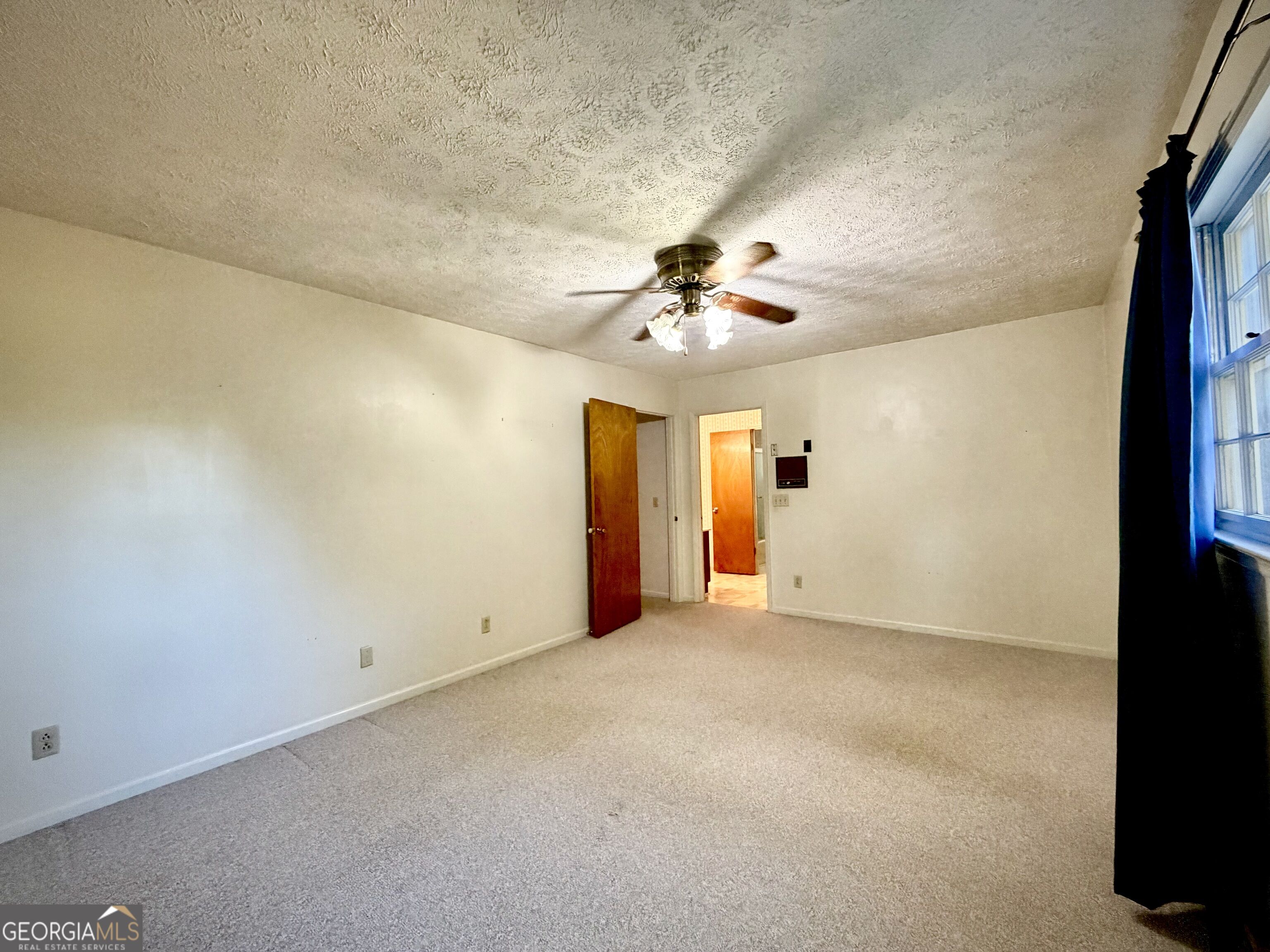 Empty carpeted room with a ceiling fan, two open wooden doors, and a curtained window.