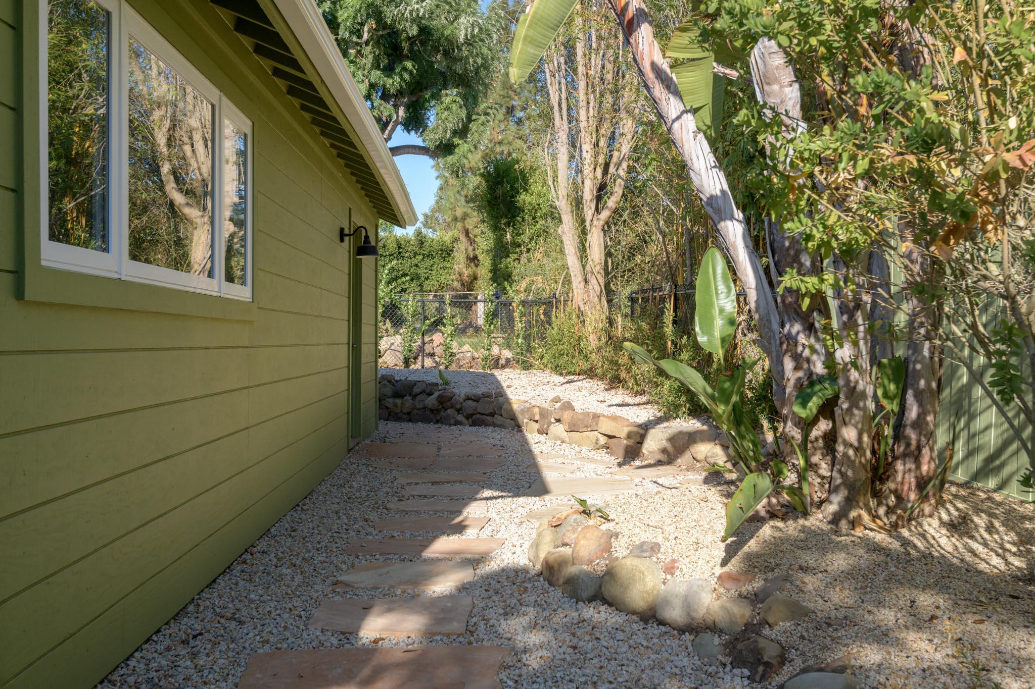 Gravel path with stepping stones beside a green-sided home, surrounded by dense trees and tropical plants.