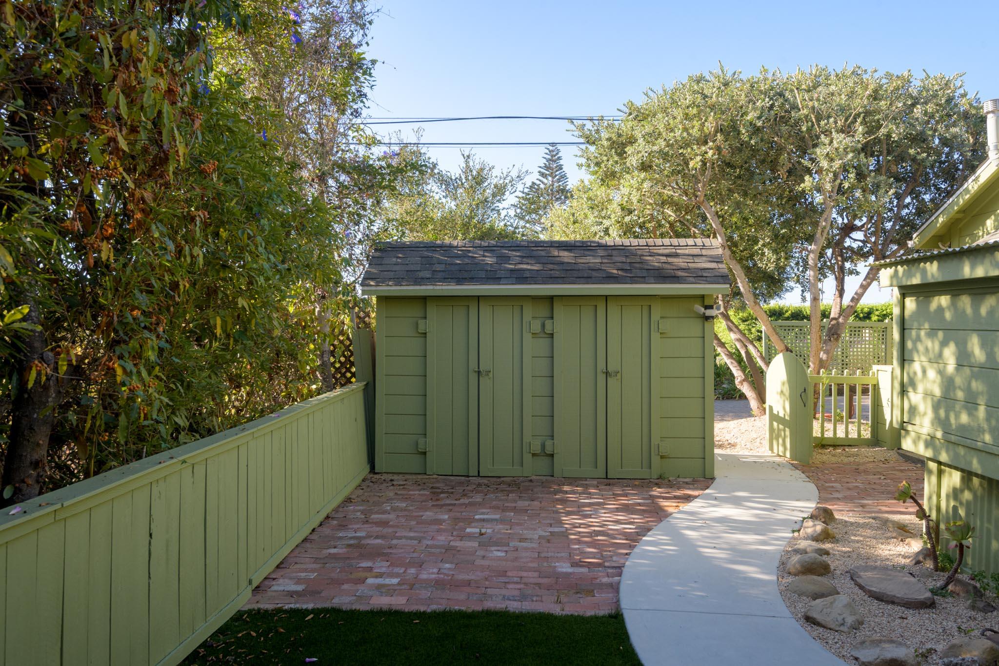 Green garden shed at the end of a brick path, beside a curved walkway and mature trees.