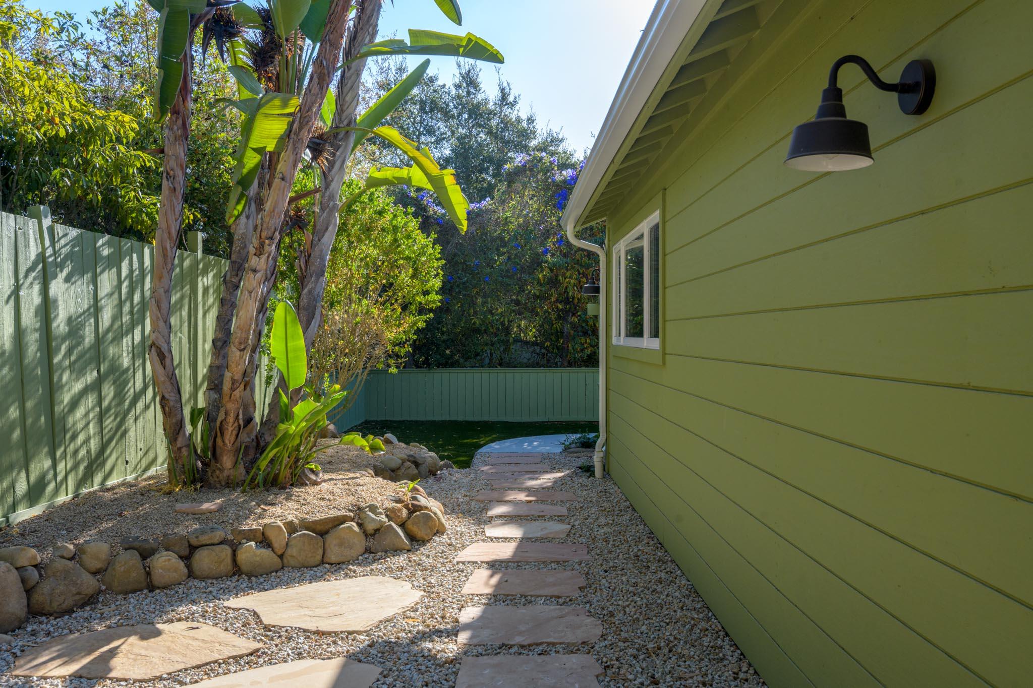 Landscaped side yard with gravel paths, tropical plants, and a small water feature beside a green house.