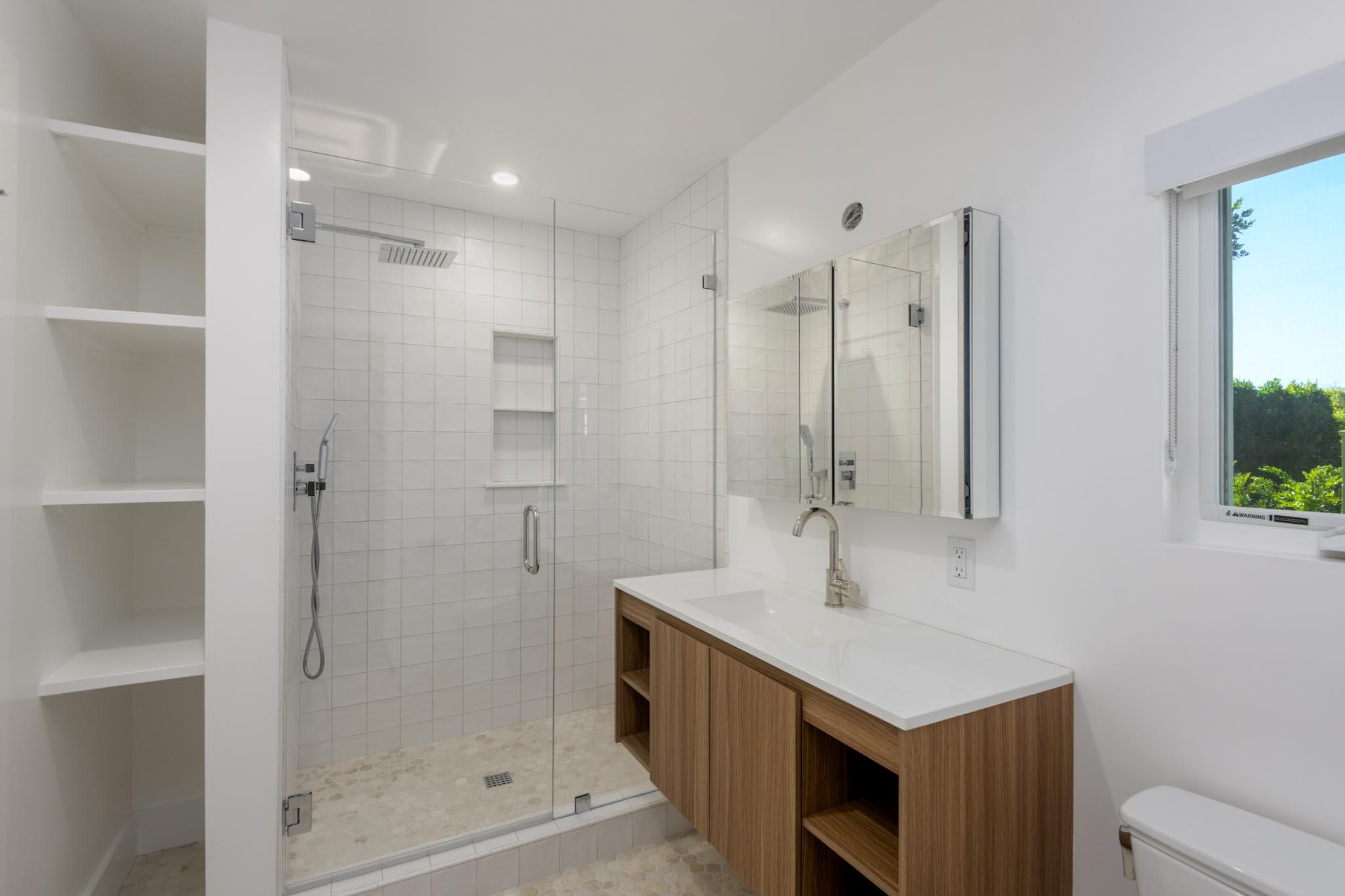 Modern bathroom with a glass walk-in shower, wood vanity, built-in shelves, and a window overlooking greenery.
