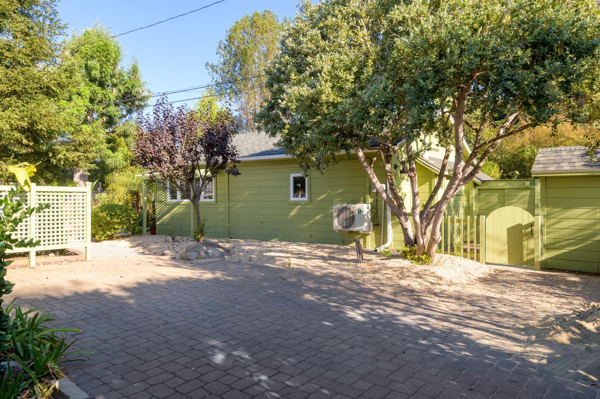 Green-sided outbuilding beside a paved courtyard, shaded by mature trees and enclosed by a gate.