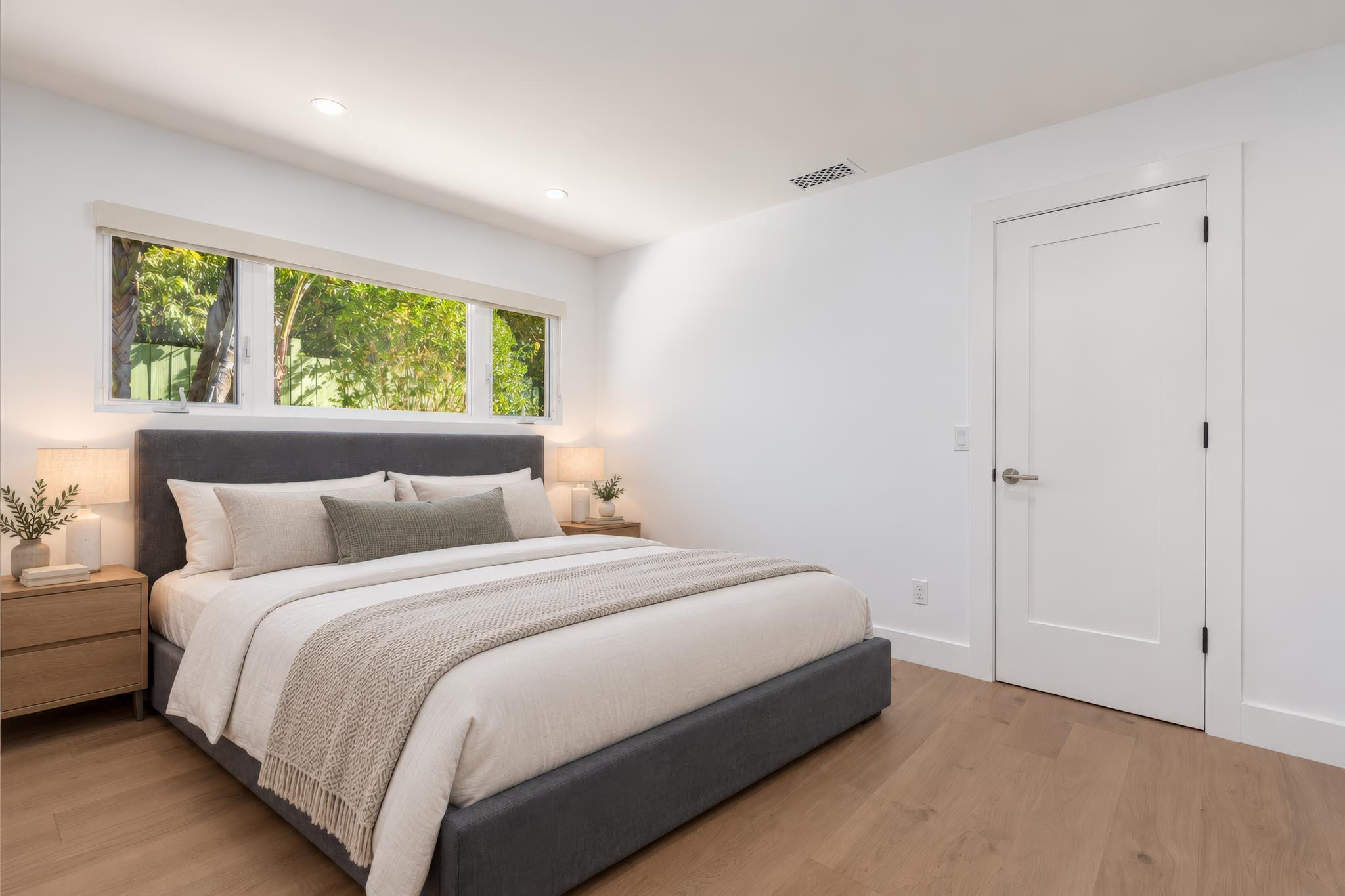 Staged bedroom with a gray upholstered bed, white bedding, and a window overlooking greenery.