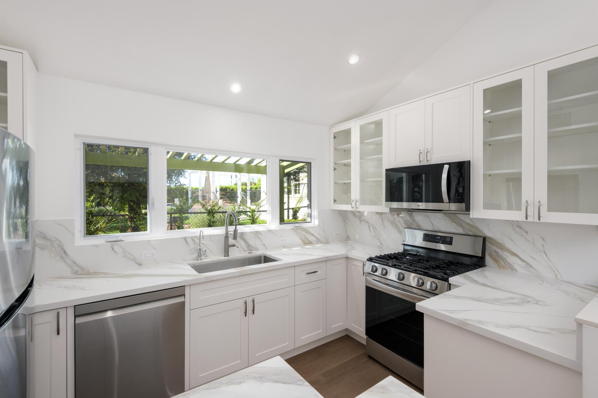 Bright white kitchen with marble-patterned counters, stainless steel appliances, and a wide window overlooking greenery.