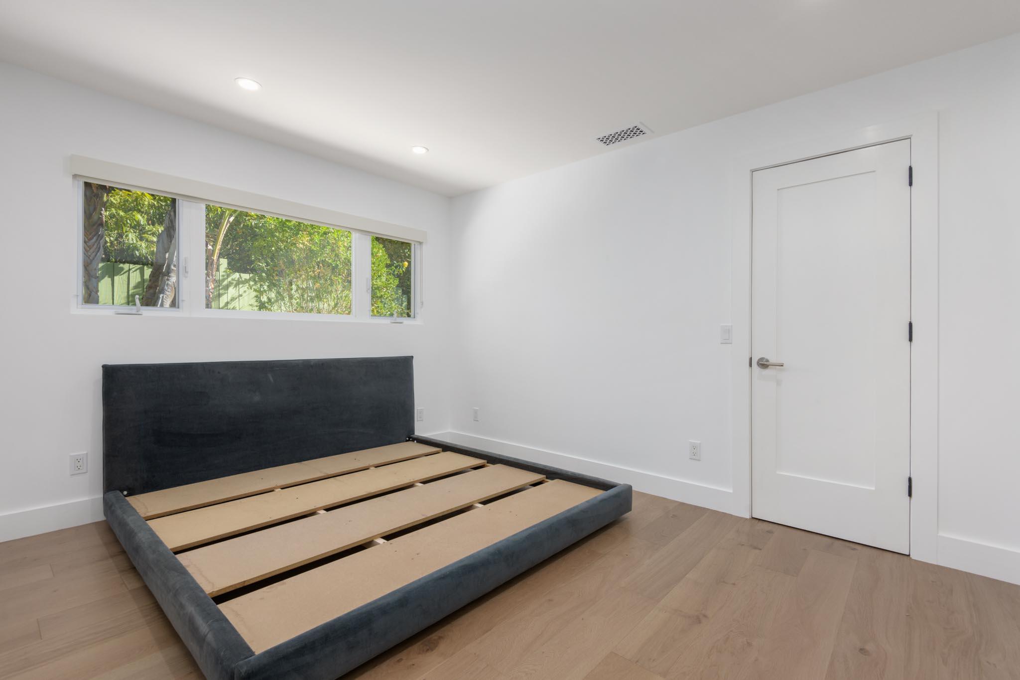 Bedroom with an unmade bed frame, wood slats, and a wide window overlooking greenery.