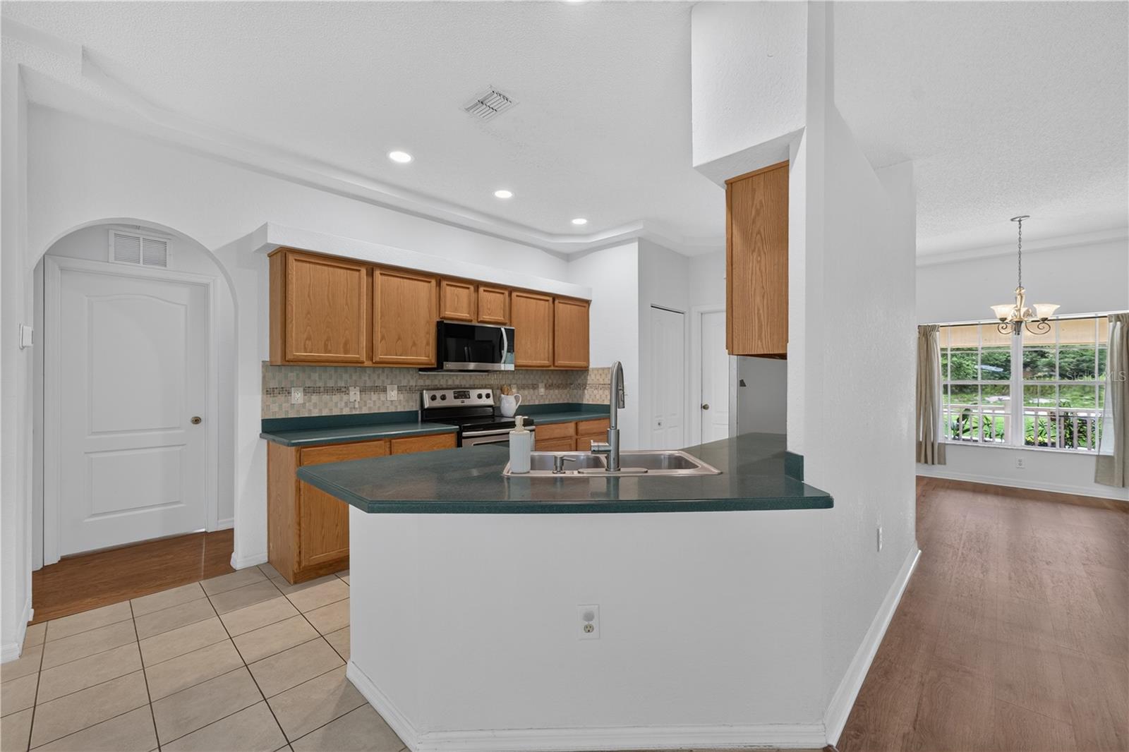 Open kitchen with wood cabinets and a green-countertop peninsula, looking into a bright dining area.