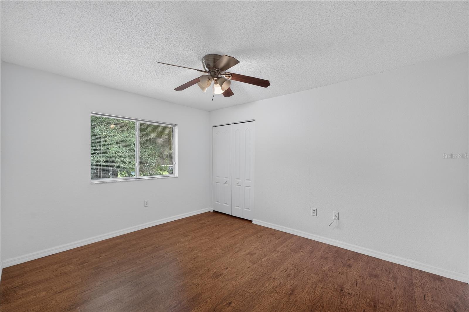 Empty bedroom with wood flooring, a window with blinds, double closet doors, and a ceiling fan.